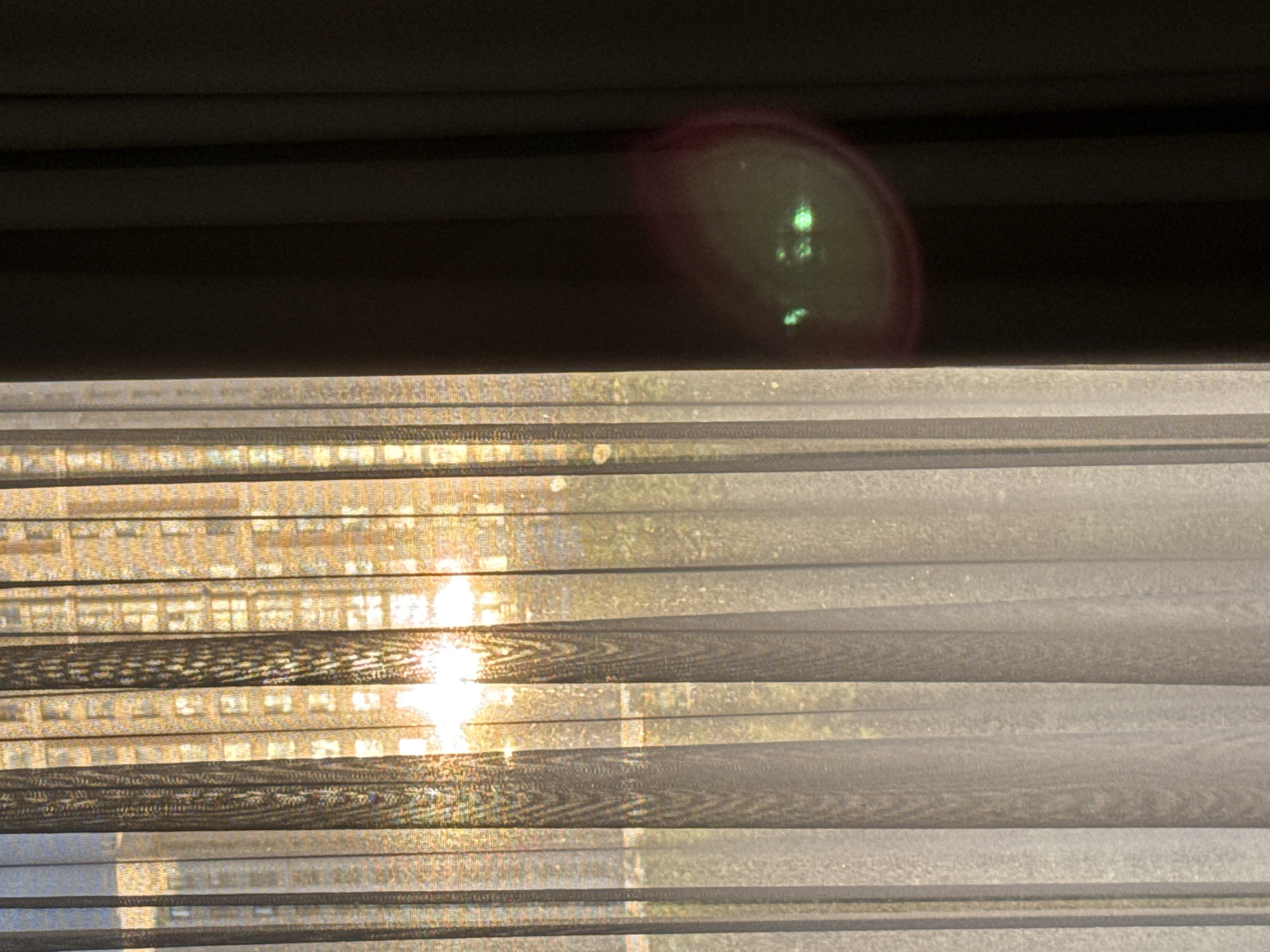The image shows a window with partially drawn blinds. Outside, bright sunlight is visible through the slats of the blinds. A dark, undefined area occupies the top portion of the frame, possibly a window frame or another structure. There is a lens flare in the upper right portion of the frame. I have no information about Khimki, Russia.