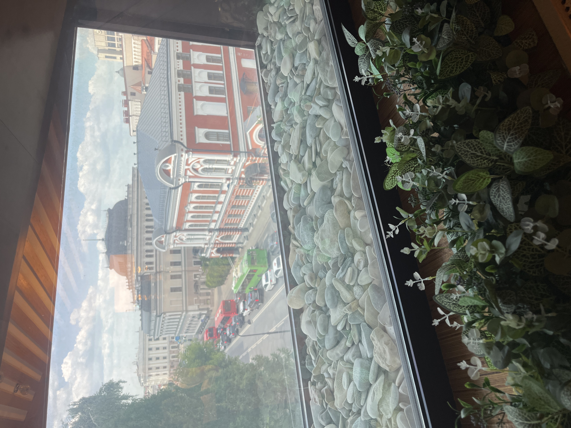 This is a photograph taken from an indoor vantage point, looking out a window that overlooks a city street. In the foreground, a decorative arrangement of various sizes and colors of smooth, light-colored stones fills the bottom portion of the window sill. Beyond the stones, a street scene unfolds with several vehicles, including red buses and cars, traveling in traffic. The buildings across the street are predominantly historic and ornate, with red and white facades, classical architectural details, and prominent rooflines. The sky above is partly cloudy with a mix of blue sky and white clouds. The overall impression is of a view from a building with a design aesthetic that incorporates natural elements like stones and plants, looking out onto a bustling urban environment with significant historical architecture. The city of Kazan, Russia is visible in the background, specifically the Kazan Kremlin complex, with its distinctive domes and architecture.