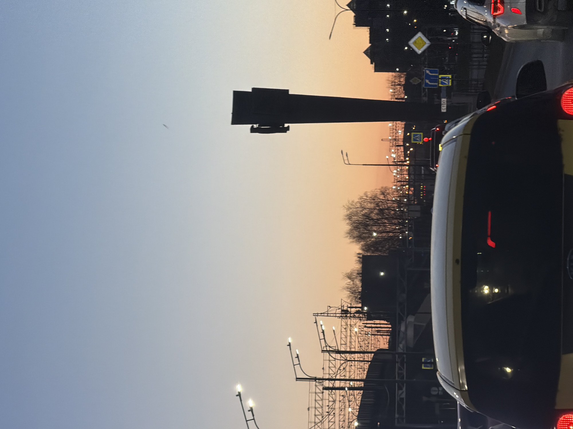 The image depicts a street scene at dusk in Perm, Russia. The sky transitions from blue to orange. A vehicle, partially visible on the right side of the frame, has a yellow stripe. Infrastructure includes overhead power lines, streetlights, and a prominent dark, elevated structure. Several road signs are visible. Bare trees are silhouetted against the horizon.