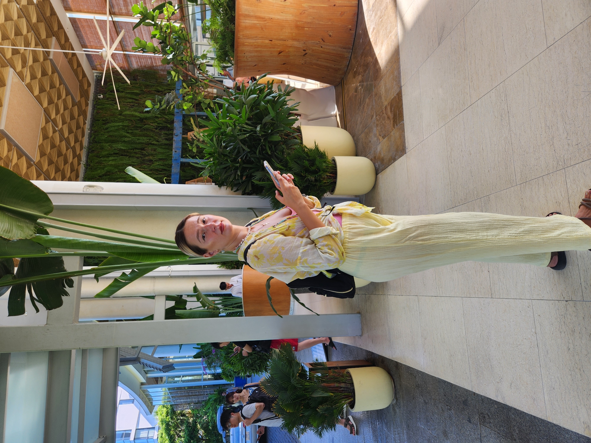 A woman stands in an outdoor covered walkway with large tropical plants and light-colored tile flooring. She is wearing a light yellow, patterned long-sleeved shirt over a pink top, and loose-fitting pale yellow pants. A black backpack is visible. She is holding a smartphone in her right hand. In the background, several other people are walking, and a man in a white shirt is standing behind a wooden reception desk. The location is identified as Da Nang, Vietnam, suggesting a hotel or resort environment with lush greenery and modern architecture.