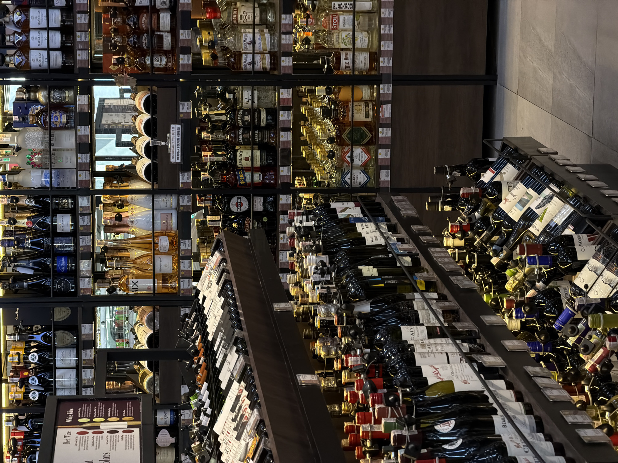 The image depicts the interior of a liquor store, possibly located in North Kuta, Indonesia, as indicated by the sign "YOU BREAK IT, YOU BUY IT MERUSAK BERARTI MEMBELI." The store is well-stocked with a wide variety of alcoholic beverages, including wine, whiskey, vodka, and other liquors. Bottles are displayed on shelves and racks, with the wine section prominently featured in the foreground. The store appears to be well-lit, with a clean and organized layout. No people are visible in the image.
