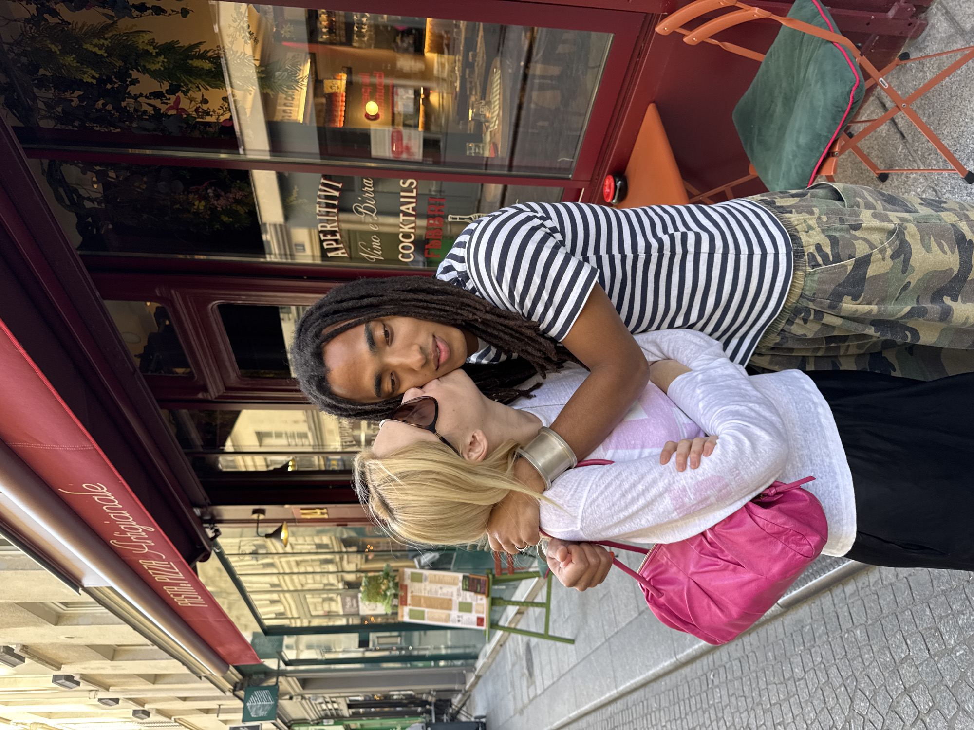 An outdoor shot depicts two individuals embracing affectionately in front of a café or restaurant, likely in Paris, France. The image is rotated 90 degrees counter-clockwise, showing the scene as if viewed from above.
The person on the left (when the image is upright) has dark dreadlocks and is wearing a black and white striped short-sleeved t-shirt paired with camouflage cargo pants. They are looking gently towards the camera. Their left arm is around the second person. A silver cuff bracelet is visible on their wrist.
The second person, embraced by the first, has blonde hair and sunglasses pushed up onto their head. They are wearing a white long-sleeved shirt, possibly with a faint pink design on the front, and dark pants. Their eyes appear closed or mostly closed as they lean into the shoulder of the first person, holding onto them. A bright hot pink handbag hangs from their arm.
The background reveals the exterior of an establishment with large glass windows and a reddish-brown facade. Reflected in the windows are nearby buildings and the street. Visible text on the window includes "APERITIVI", "Vino e Birra", and "COCKTAILS", indicating it's a place serving drinks. Above the window, a deep red awning extends, bearing partially visible white script that reads "PASTA PIZZA Artigianale". Outdoor seating, including a chair with a green cushion, is visible to the right of the couple. The ground is paved with stone blocks. The lighting suggests it is daytime, with a soft, warm glow. The overall mood conveyed is one of affection and connection between the two people.