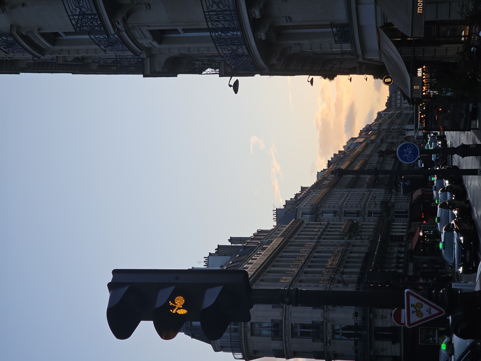 The image depicts a street scene in Paris, France. The perspective is looking upward from street level, capturing the characteristic Haussmannian architecture of the city, with ornate buildings lining both sides of the street. The sky is visible between the buildings, displaying a soft gradient of color from light blue to a warm orange hue, indicating either sunrise or sunset. A traffic light with a pedestrian crossing signal, showing a walking figure lit in yellow, is in the foreground. In the background, vehicles including taxis are parked along the street, and various road signs are visible. A storefront awning is present on the right, with the name "BRIA" visible in the signage. A bicycle lane symbol is mounted on a post to the right.