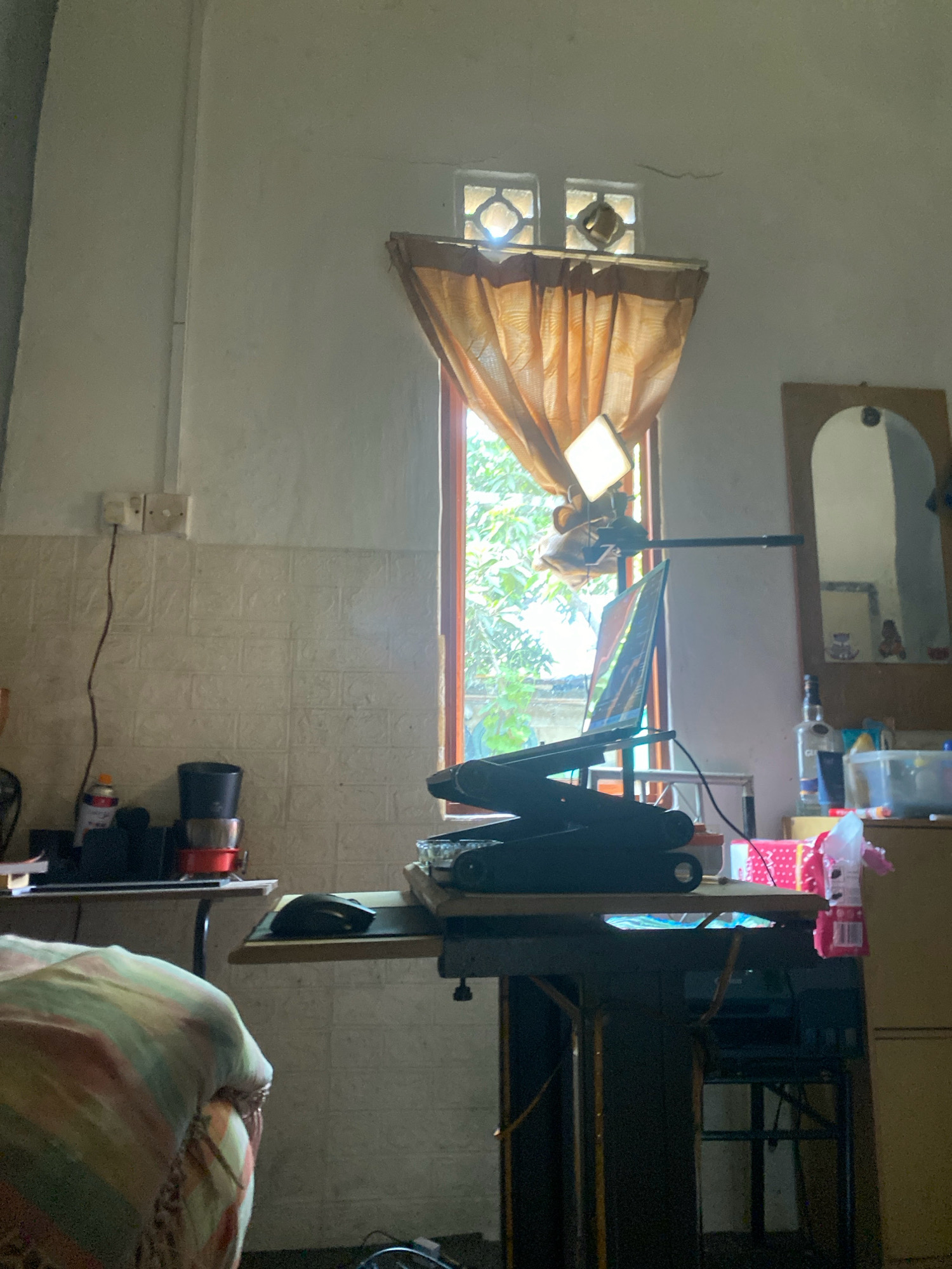 The image depicts an indoor scene, likely a residential room in Batam, Indonesia. The room appears to be a workspace or study area, with a desk set up near a window. On the desk, a laptop is open, supported by a stand, and a computer mouse is visible. A lamp is positioned above the laptop, casting light. The window, covered by an orange curtain, reveals greenery and trees outside, suggesting a tropical environment. The room's walls are painted white, with some textured tiling in the lower section near the desk. Electrical outlets are mounted on the wall. Various objects are scattered around the desk and surrounding areas, including bottles, containers, and what appears to be a sewing machine base underneath the desk. The overall impression is of a functional, albeit somewhat cluttered, living space.