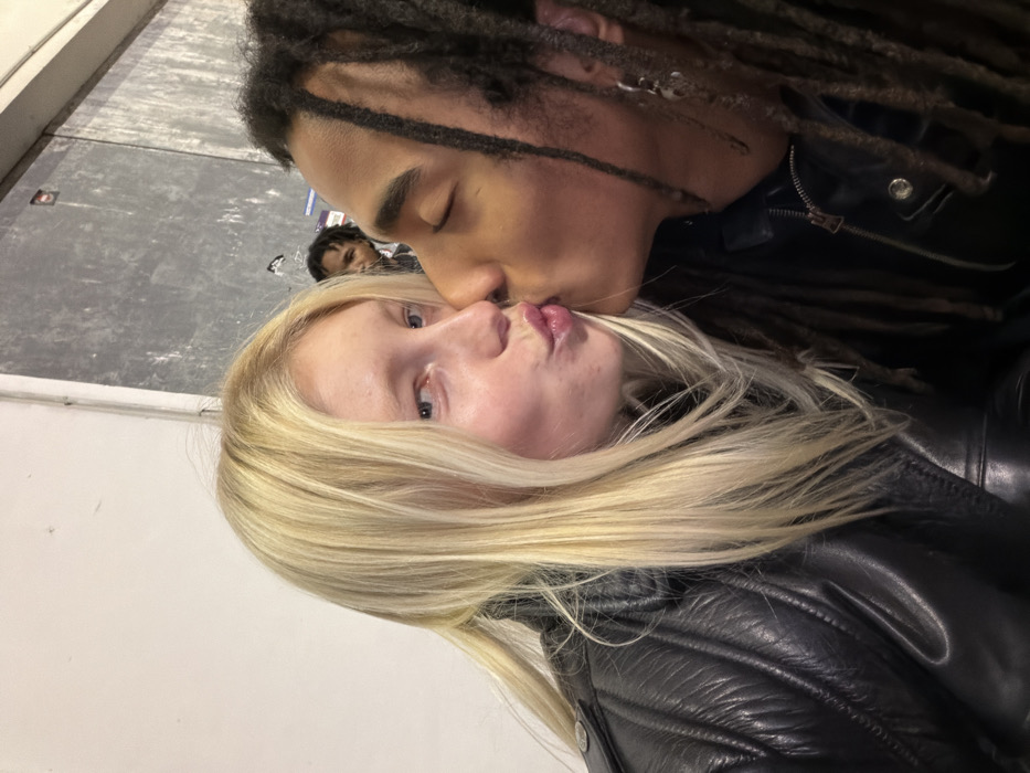 In Bagnolet, France, two individuals are depicted in a close-up, chest-level portrait. A female with long blonde hair and fair skin is on the left, looking directly at the camera with blue eyes and pursed lips. She wears a black leather jacket with quilted detailing on the shoulder. Her lips are pressed against the lips of a male on the right, who has a darker skin tone, dark dreadlocks, and closed eyes. He wears a dark zippered jacket, also appearing to be leather, and a silver earring with a small charm is visible on his left ear. The background features a plain white wall on the left and a darker, textured grey wall with some markings on the right. A third individual with dark skin and dark hair is partially visible in the upper background, positioned between the heads of the two main subjects, looking towards the right side of the frame.