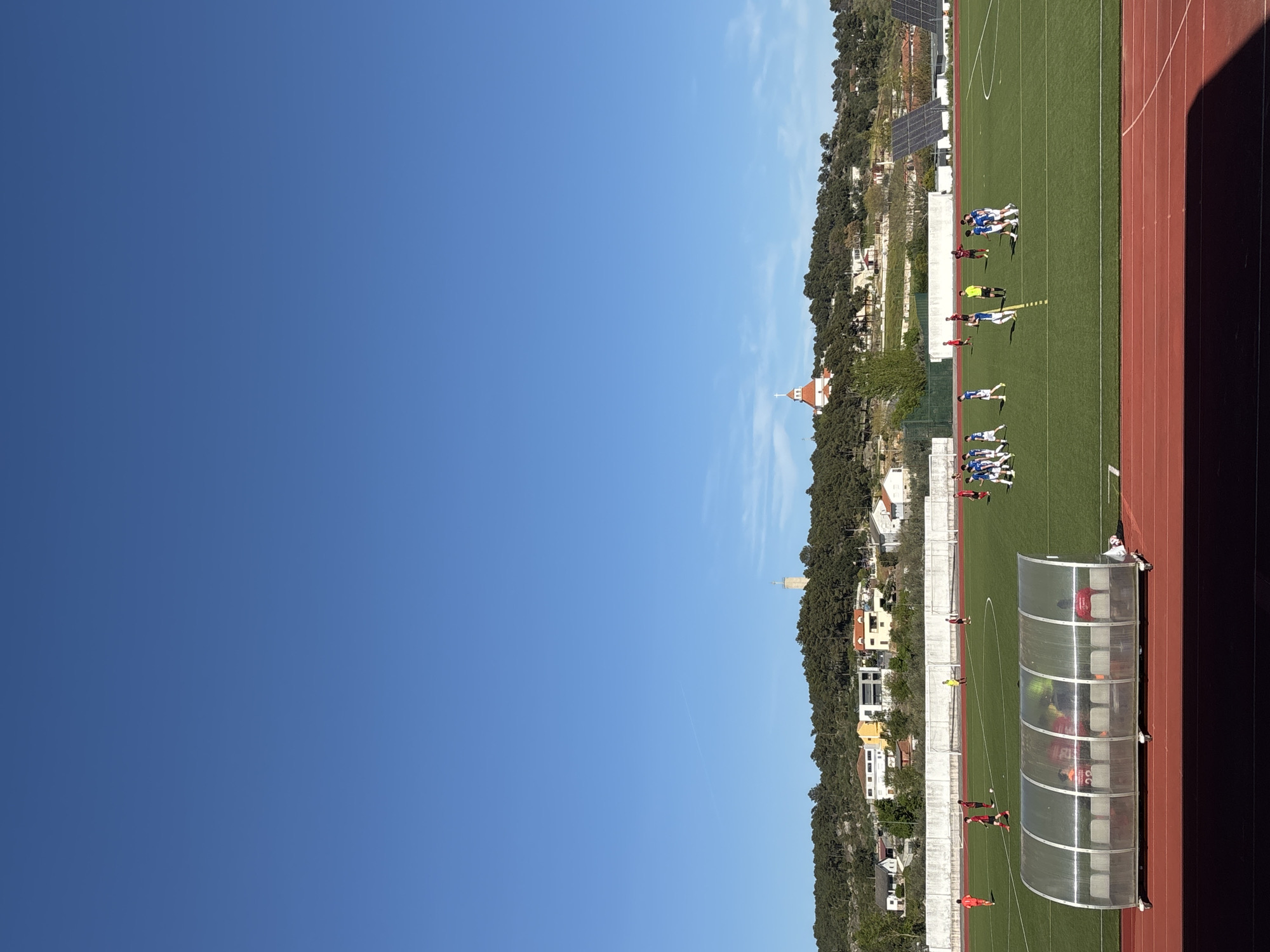 The image depicts a soccer match in progress on a green field, alongside a red running track, under a clear blue sky. A hillside with houses, trees, and structures, including a tower, flanks the field. On the sideline, a team dugout is visible. Several soccer players in blue and white uniforms are grouped on the field, suggesting a potential play or set piece. The location is likely Valpaços, Portugal, based on the provided contextual information.