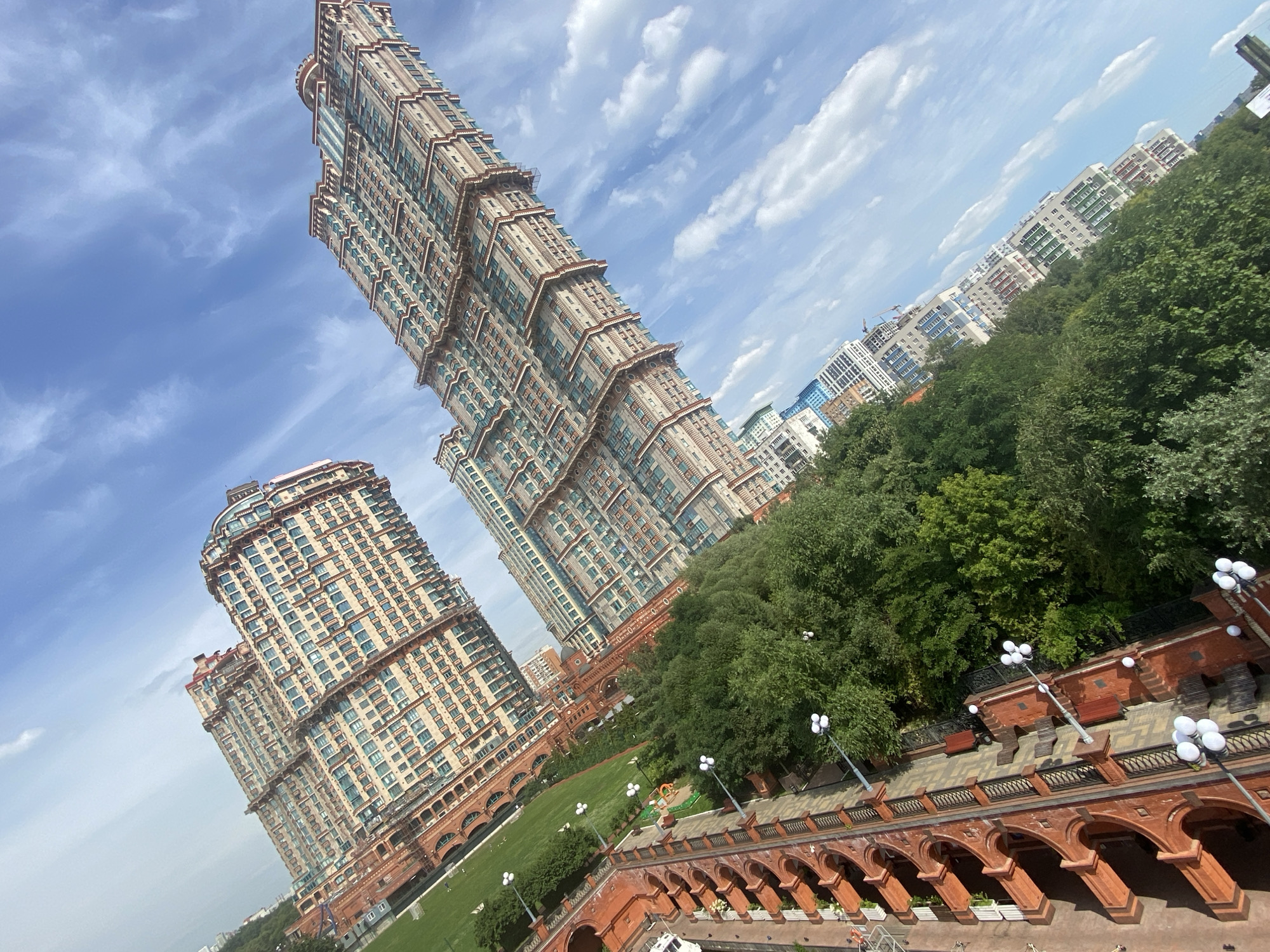 The image shows a tilt-shifted view of a city skyline featuring several large, modern apartment buildings. The buildings are tall and have many windows, with a predominantly beige and red facade on the two most prominent structures. The buildings are angled diagonally in the frame, as is the sky, which is blue with scattered white clouds. In the foreground, there is a lush green park with mature trees lining a pathway. Some streetlights with multiple bulbs are visible along the pathway, and there is a red brick structure with arched openings on the left side of the frame. The scene appears to be an urban residential area with ample green space.