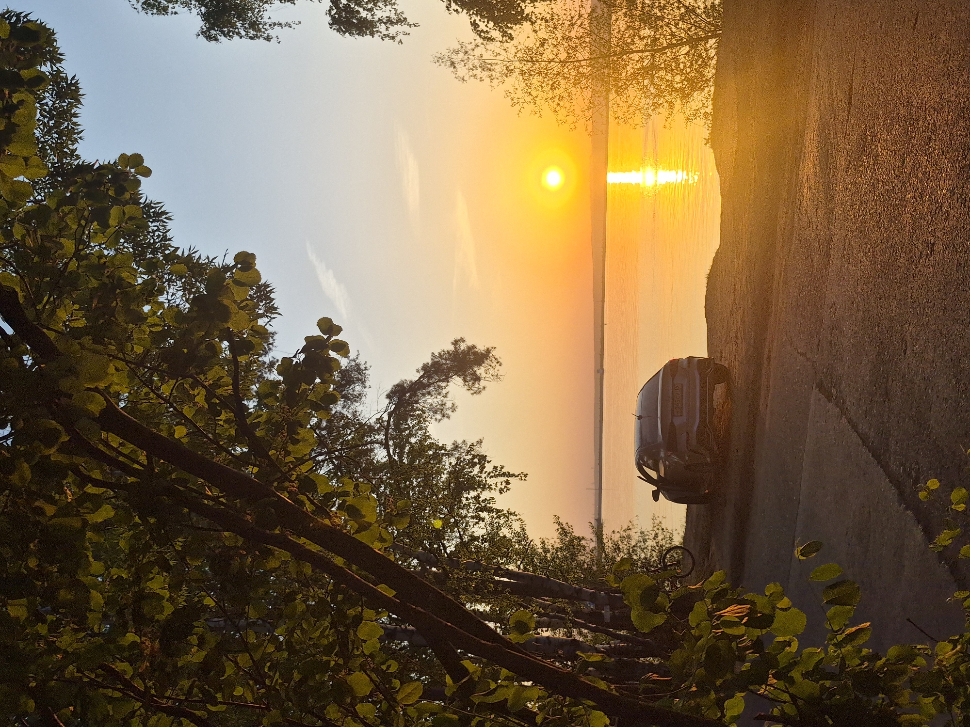 The image shows a sunset scene in an unknown location. The sun is setting over water, creating a bright reflection. A car is parked on a road next to the water. Trees are present in the foreground.