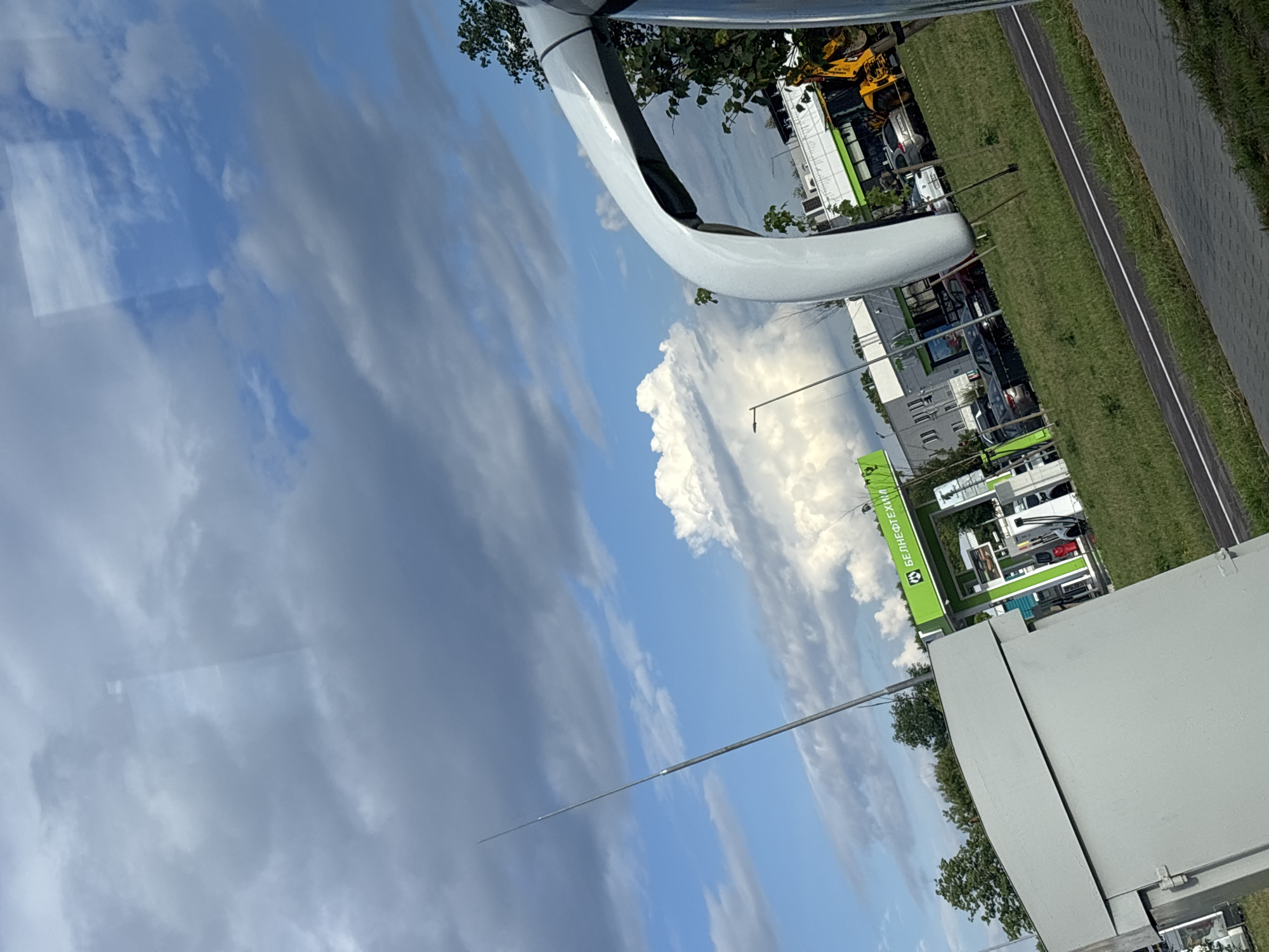 The media file is a photograph taken outdoors, featuring a bright sky with scattered clouds. The image is oriented vertically. In the foreground, a portion of a large, white, curved object is visible, possibly part of a vehicle or a structure.
The background reveals a street or road with a grassy verge. Beyond that, there are buildings, some with green awnings or signage. A yellow construction vehicle is partially visible among the buildings. Trees are scattered throughout the scene.
A prominent green sign with white lettering is visible. The text on the sign appears to be in Cyrillic script and reads "БЕНЕКОТЕХНИК" and below it, "ВАСИЛЕВ".
The lighting suggests it is daytime, likely a sunny day given the brightness of the sky and clouds. The weather appears to be clear with some cloud cover. The location context provided indicates the setting is within the Senitsa village Council in Belarus. There are no people clearly visible in the image. The overall scene depicts an outdoor commercial or industrial area.