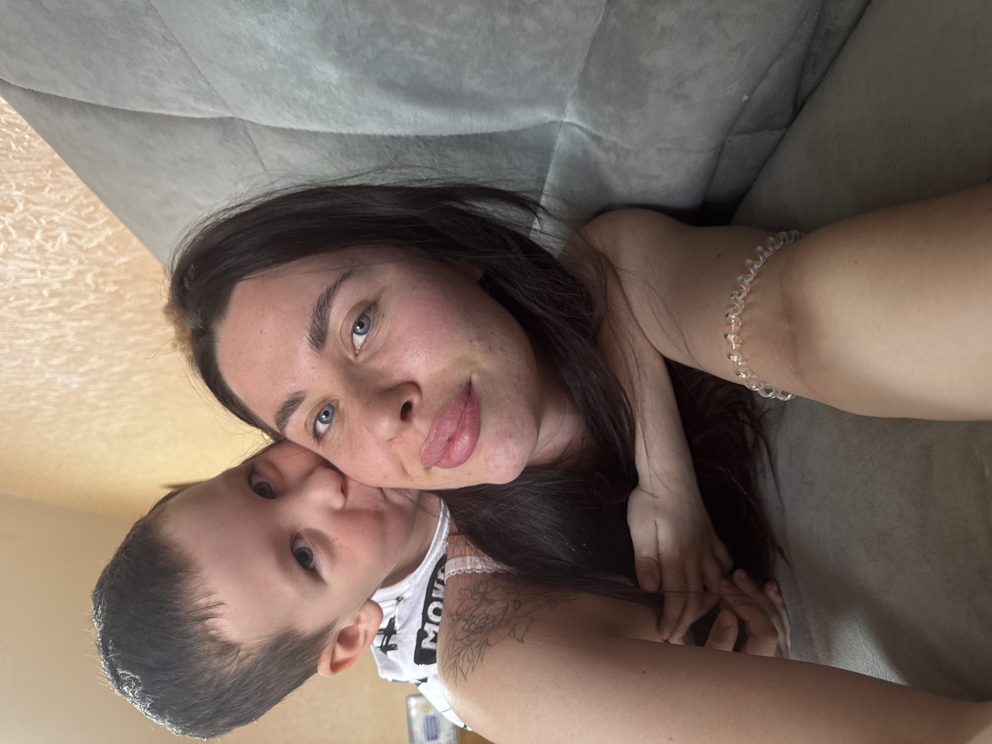 A woman with long, dark hair and blue eyes lies on a grey sofa. She is positioned in a way that suggests she is taking a selfie. A child with short, dark hair and similar eyes is nestled close to her. The child's face is partially obscured. The child wears a white tank top. A bracelet is visible on the woman's wrist. A tattoo is present on the woman's shoulder. The background includes a light-colored wall. The location is Chaikovsky, Russia.