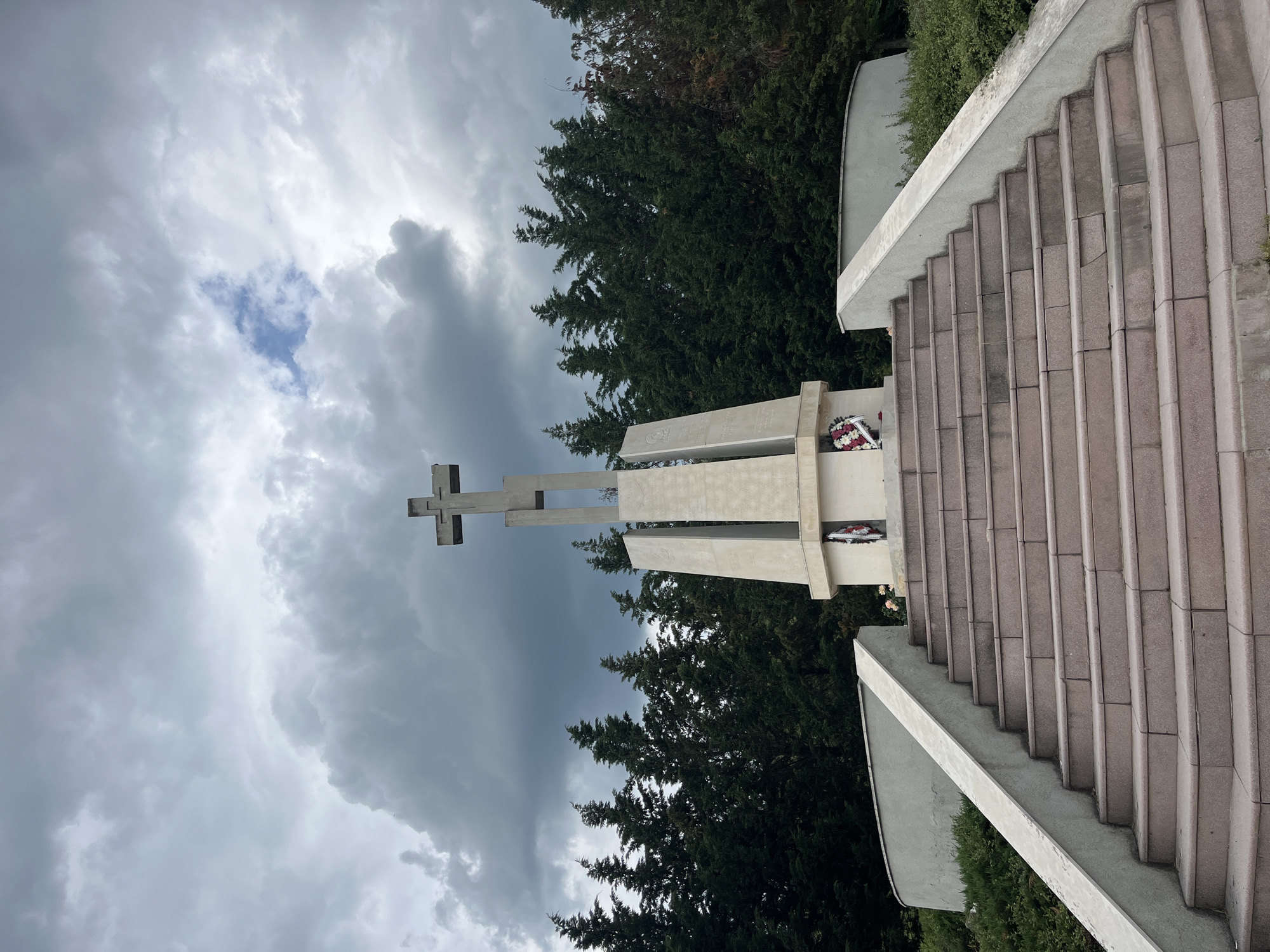 The image shows the Monument of Bulgarian-Soviet Friendship in Varna, Bulgaria. The monument consists of a large concrete structure with a prominent cross at the top. Below the cross are rectangular concrete slabs with wreaths attached. To the right of the monument are a set of stone stairs leading up to the structure. The background features dense green trees and a cloudy sky. The image appears to be taken from a low angle, looking up at the monument. There are no people visible.