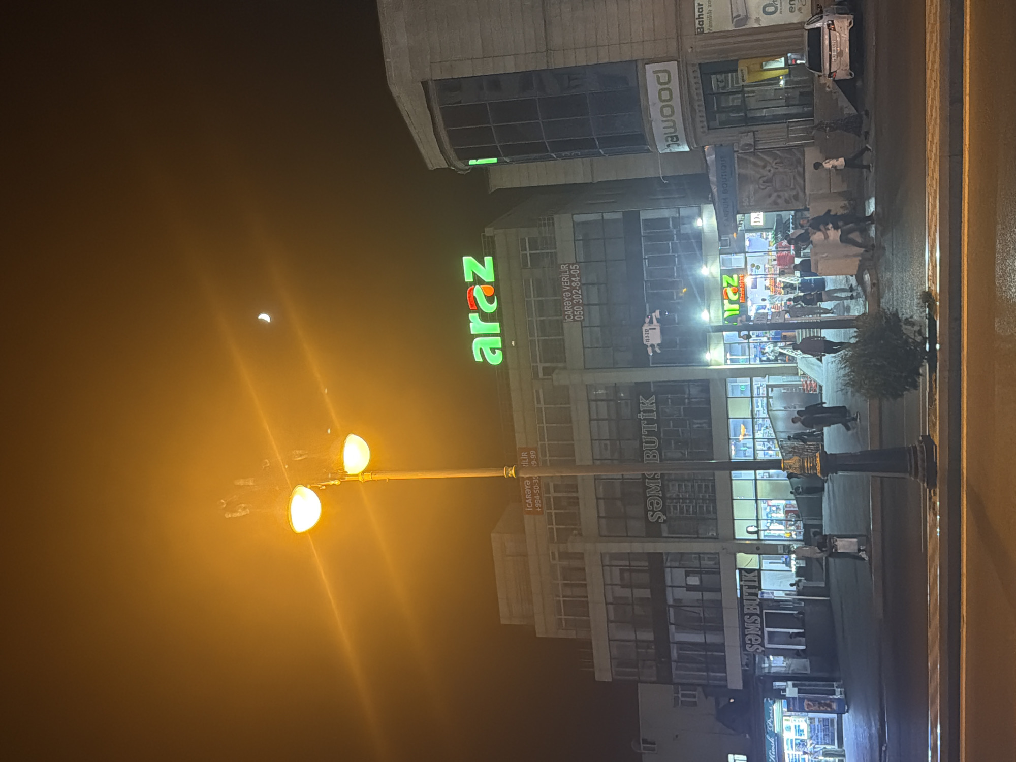 In an outdoor urban setting at night, a street lamp with two large lights illuminates the foreground, casting a warm glow. A crescent moon is visible in the dark sky. Several buildings line a street, some with illuminated storefronts. One building prominently displays a green and orange "araz" logo. Other signs, such as "SƏMS BUTİK" and "PARFUM BOUTIQUE," are also visible. People are walking on the sidewalk and crossing the street, some in small groups. A white car is parked on the right side of the street. The ground is paved with asphalt and concrete.
