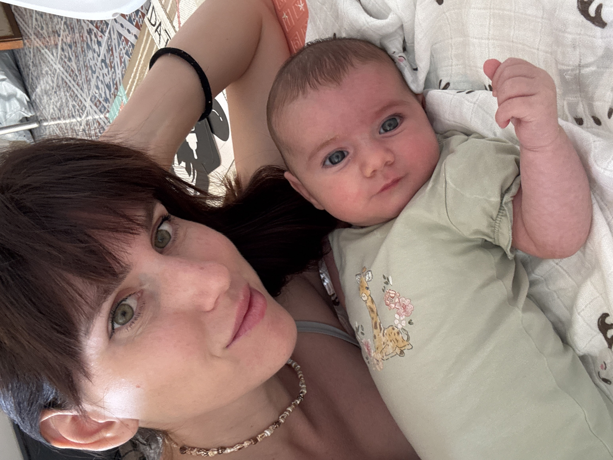 The image shows a woman and a baby lying down. The woman is in the foreground, with her face close to the camera. She has dark hair and green eyes. She is wearing a necklace. The baby is lying next to her, with its head resting on a patterned blanket. The baby has blue eyes and is wearing a light green onesie with a giraffe and floral print. The background is blurred but appears to be an indoor setting with a patterned rug and some furniture. The image is taken from a close-up perspective, focusing on the faces of the woman and the baby. There is no discernible indication of the location in the city of Isola delle Femmine, Italy.