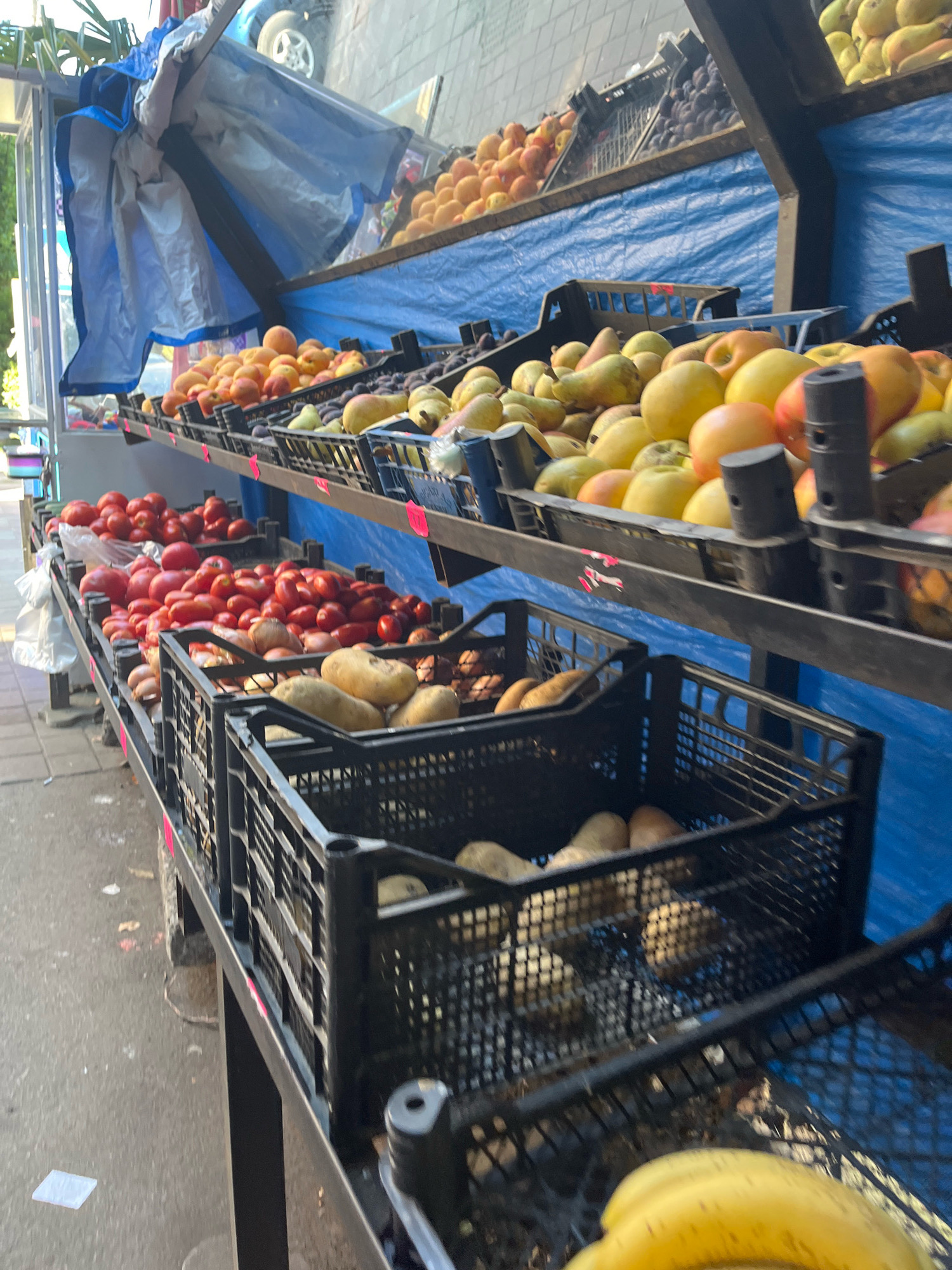 A close-up, eye-level, vertical shot captures an outdoor fruit and vegetable stand in Kobuleti, Georgia. The foreground is dominated by a black plastic crate filled with yellow bananas. Behind this, a black crate contains potatoes. Further back, more black crates are visible, holding an assortment of produce. Two long shelves, covered by a blue tarp, are laden with various fruits. The upper shelf displays peaches and plums, while the lower shelf features an abundance of red tomatoes. A hint of a vehicle and greenery suggests an urban setting. The overall scene depicts a marketplace stall displaying fresh produce.