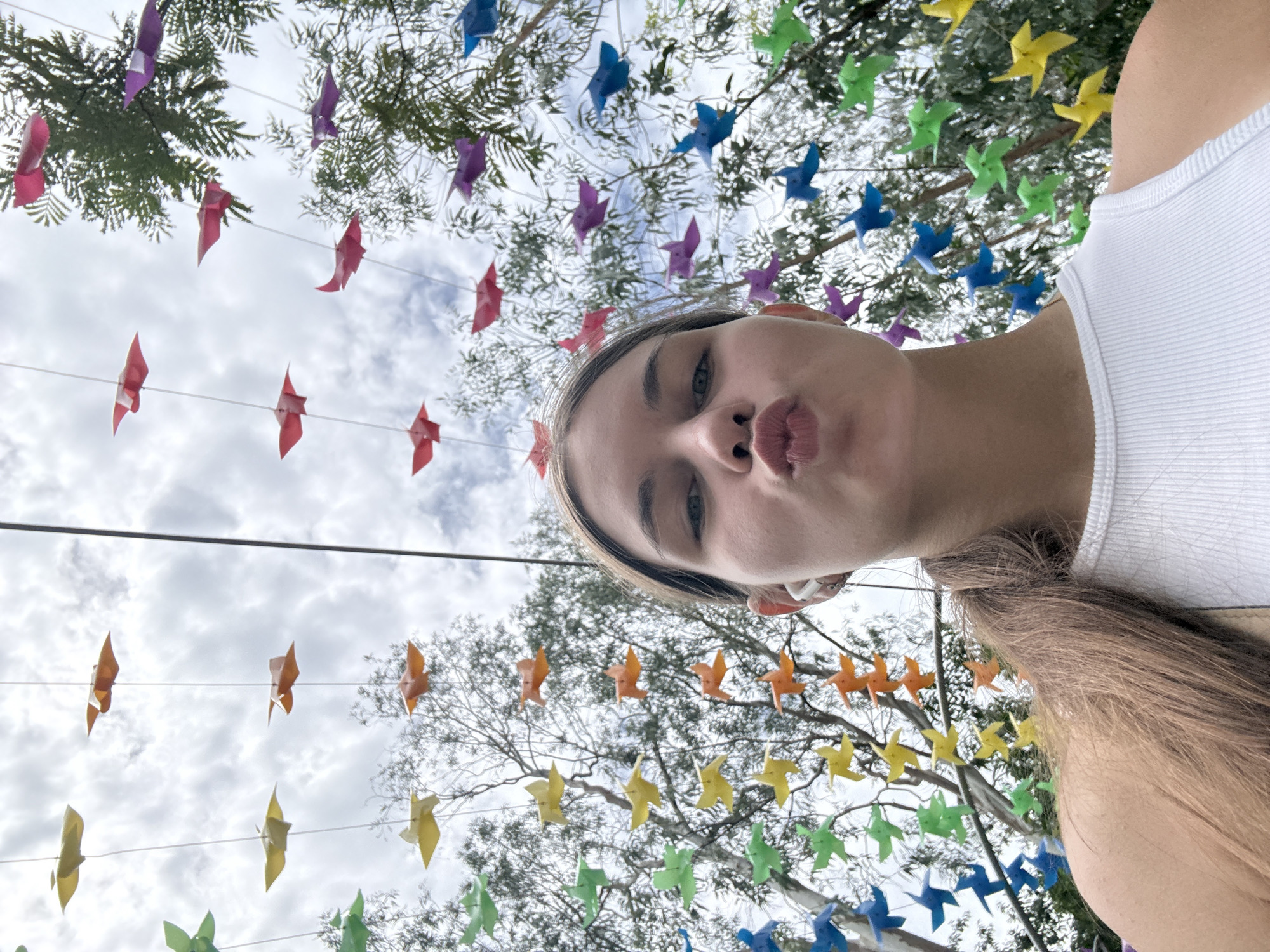 The image shows a young Caucasian woman with long brown hair pulled back in a ponytail, wearing a white tank top and wireless earbuds. She is leaning into the camera with her lips pursed. The background is an outdoor scene with trees and a cloudy sky. Hanging from strings are numerous colorful origami birds in various colors, including green, yellow, orange, red, pink, purple, and blue. The origami birds are arranged in garlands, creating a decorative canopy overhead. The overall impression is festive and cheerful. The context of Nha Trang, Vietnam, suggests this might be a public art installation or decoration in a park, street, or tourist area.