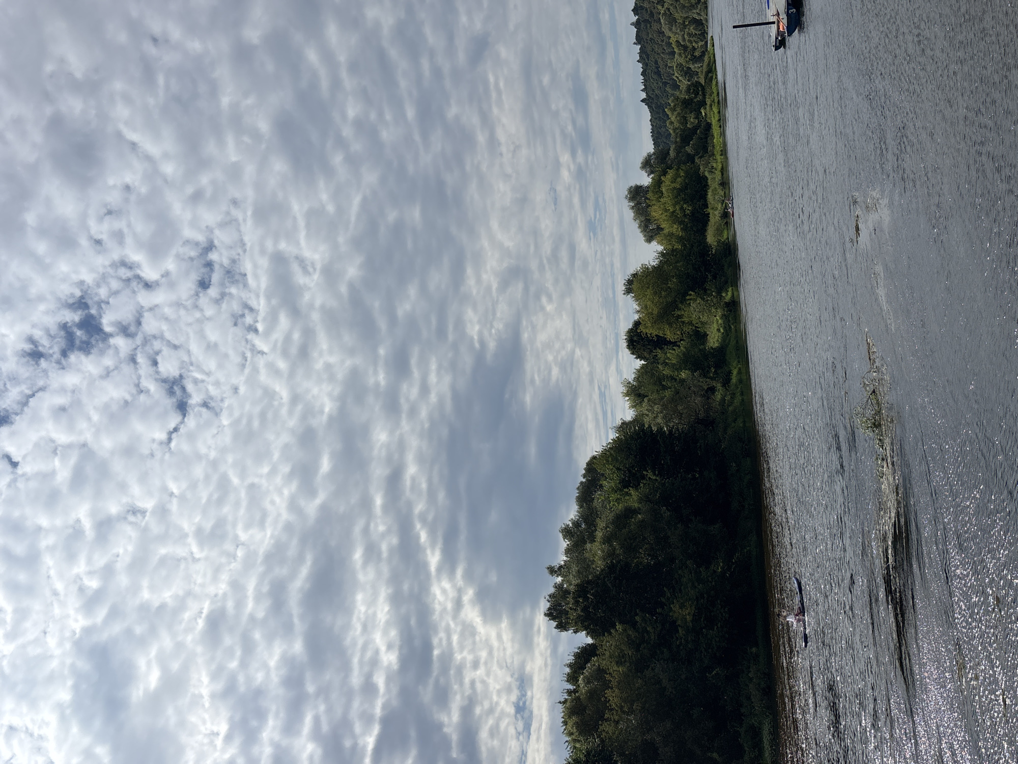 The image shows a wide river with trees lining the banks. The sky is filled with dramatic, cloudy formations. In the foreground of the river, a person is seen paddleboarding on a calm, rippling surface that reflects the light. Further out in the river, two small boats are visible. The distant bank is covered with a dense line of green trees, and beyond that, rolling hills covered in vegetation. On the right side of the river, several boats are moored to a dock. The scene depicts a natural landscape likely within or near Zvenigorod, Russia, given its characteristic river and forest scenery.