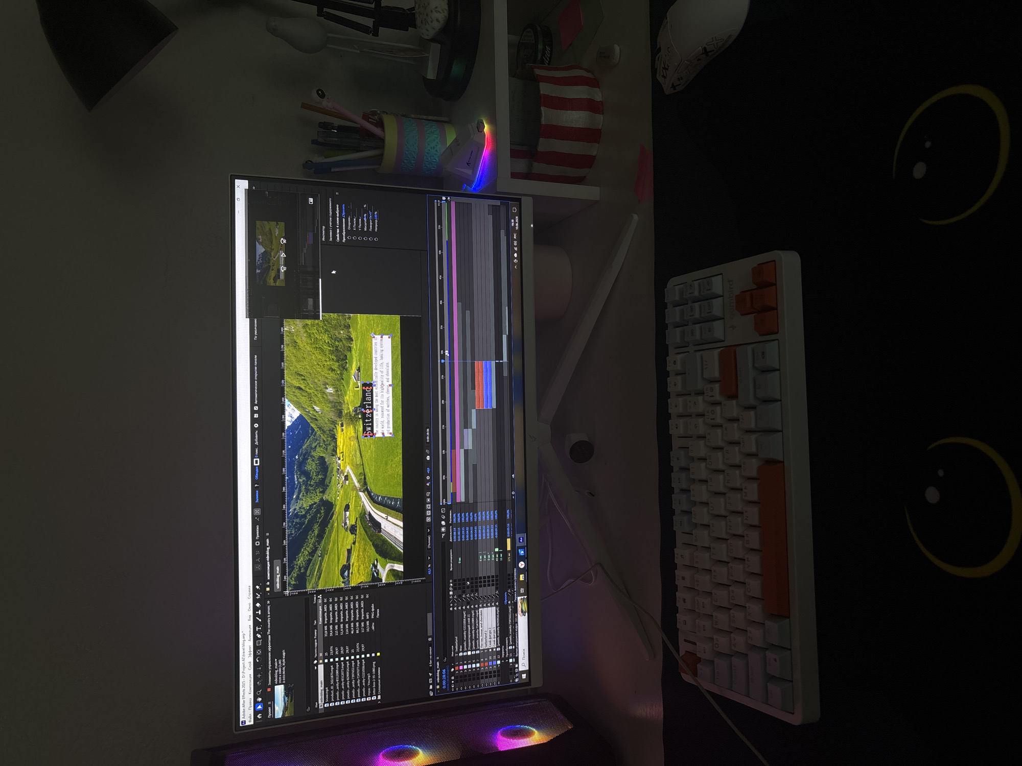 The image depicts a dimly lit desk setup, likely in a home office or gaming room. The primary focus is a large computer monitor displaying Adobe After Effects, a video editing software. The screen shows a project timeline with various video and audio tracks, a preview window with footage of a scenic landscape with the text "Switzerland" overlaid, and several other software panels.
To the left of the monitor is a computer tower with RGB lighting. To the right of the monitor, there's a shelf with several items, including a pen holder filled with pens and markers, a desk lamp, and a decorative item with red and white stripes.
In front of the monitor is a white mechanical keyboard with some orange-accented keycaps. A white mouse is positioned to the right of the keyboard on a black mousepad that features a glowing yellow smiley face design. The overall environment appears to be a personal workspace designed for creative or gaming activities.
There is no information in the image that indicates the location is in the city of Zheleznogorsk, Russia.