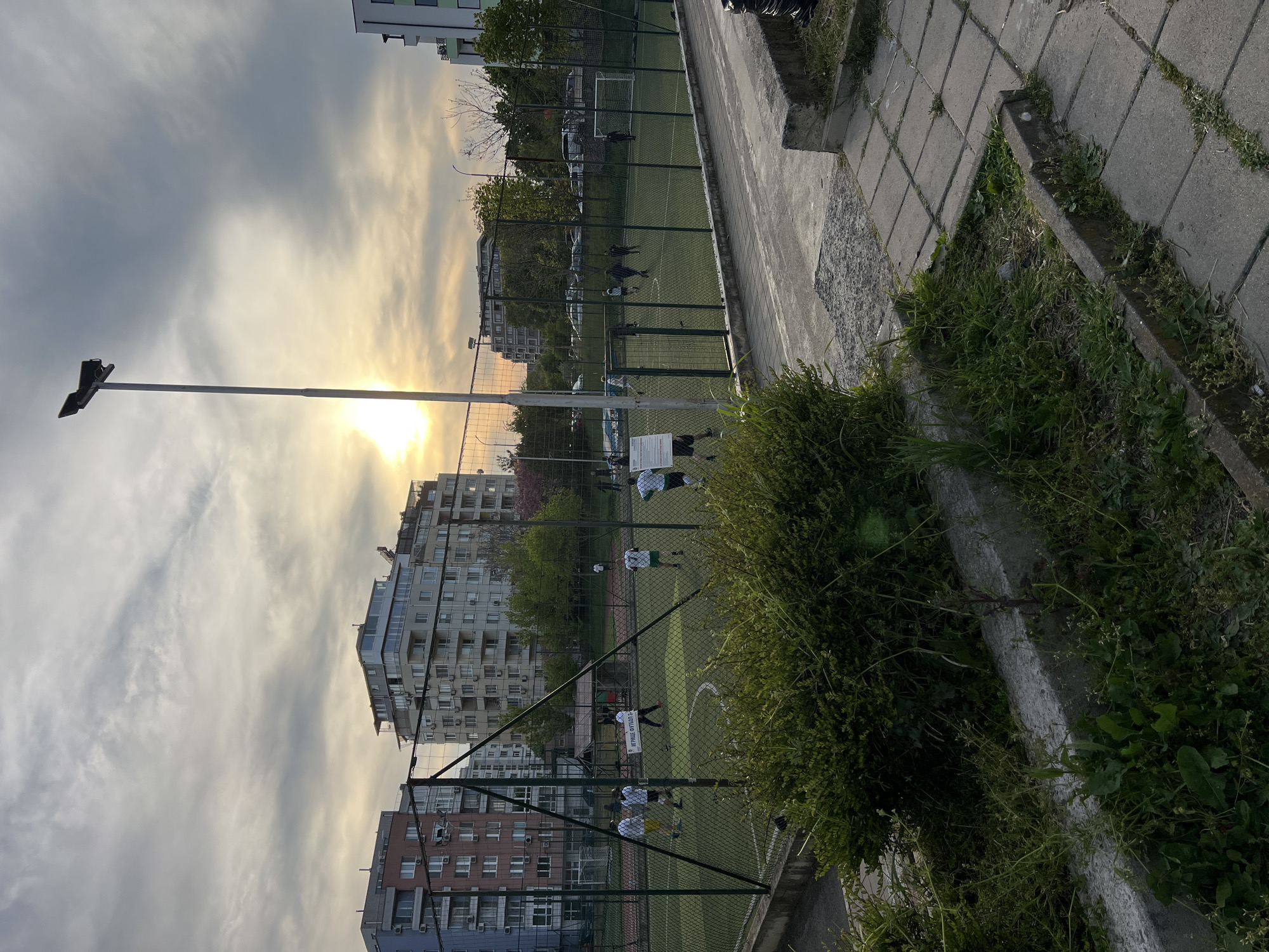 The image shows a fenced-in astroturf soccer field in what appears to be an urban setting. The field is in use, with several individuals wearing athletic clothing, seemingly engaged in a game or practice. The background consists of residential buildings, a cloudy sky with a hint of sunset, and a tall light pole extending into the upper-left. In the foreground, there is vegetation along the fence.
Based on visual cues like architectural style and the possible Cyrillic text on a sign on the field, the location is likely Burgas, Bulgaria. However, without a clearer view of the sign and precise location information, this identification is a guess.