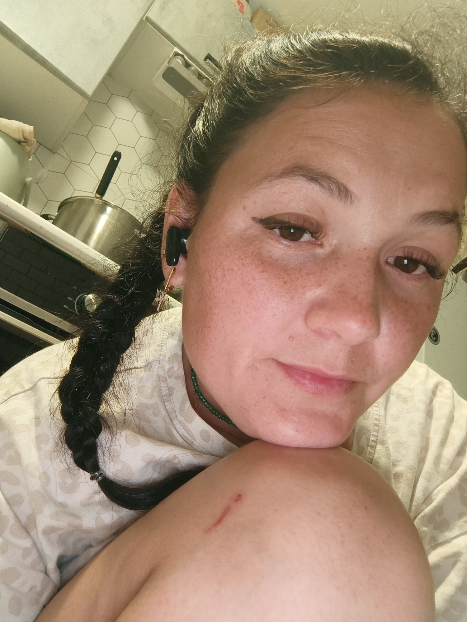 This is a close-up, selfie-style shot of a woman with brown hair styled in a braid, wearing a light-colored, patterned shirt and a green beaded necklace. She has an earbud in her left ear and is wearing a cross-shaped earring on the same side. She has freckles and what appears to be a small cut or scratch on her upper arm. The background shows a kitchen environment with white hexagonal tiles on the wall, a stainless steel pot on a stove, and kitchen cabinets. The image does not provide any specific geographical information to determine its location within the city of Chaikovsky, Russia.
