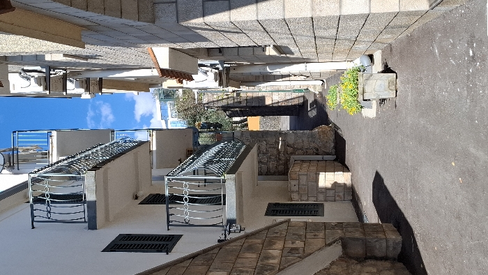 This image depicts a narrow outdoor passageway in Boreti, Montenegro, bordered by two multi-story buildings under a clear blue sky with scattered white clouds.
On the left, a building with a white stucco facade features two cantilevered balconies with dark, ornate wrought-iron railings. Below the lower balcony, a black wall lantern is affixed. Dark green louvered window shutters are visible on multiple levels. The building's base transitions to light brown stone cladding, with an angled stone structure in the foreground and a smaller, light-colored stone block structure at ground level.
On the right, a building constructed of light gray stone blocks displays several wall-mounted air conditioning units with associated piping. A segment of a terracotta-tiled roof overhang is visible. A white downspout runs vertically along this facade.
The ground surface of the passageway is dark asphalt, across which strong, elongated shadows are cast from the left-side structures, indicating direct sunlight from the right. Two rectangular, light-gray concrete planters, each containing yellow flowering plants, are positioned near the base of the right building. The passageway narrows in the background, revealing distant green foliage and a modern, light-colored building with multiple windows. No individuals or discernible activities are present within the scene.