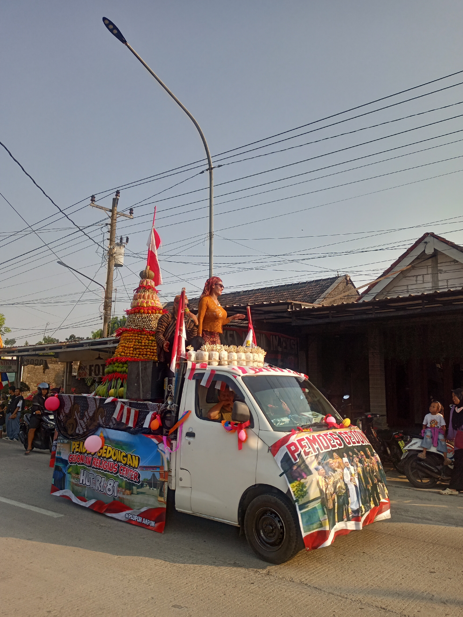 The image captures a street scene in Ngrompongan, Indonesia, during what appears to be a parade or celebration. The main focus is a white pickup truck decorated with a large banner on the front displaying a group of people in traditional attire and the text "PEMDES GEDONGAN". The truck is adorned with balloons and streamers, and on its bed, a tiered arrangement of fruits and vegetables is displayed, topped with Indonesian flags. Two individuals in traditional clothing stand on the truck, one in an orange top and the other in a brown patterned garment.
The truck is moving along a paved road, with other vehicles and pedestrians visible. To the left, a motorcycle carries a rider and passenger, and several people are walking on the side of the road. In the background, there are buildings with tiled roofs, power lines, and a street lamp. Another banner on the side of the truck reads "PENDUDUKAN GEDONGAN RELIGIUS CENTER HUT RI 81" and "#PLUPUH AMPUH". A young child is seen sitting on a motorcycle on the right side of the image, looking towards the truck. The overall atmosphere suggests a community event taking place outdoors under a clear sky.
