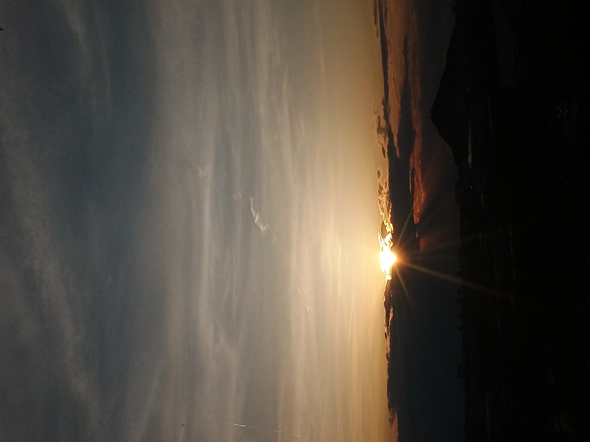 The image depicts a sunset in Plovdiv, Bulgaria. The sun is positioned near the horizon, partially obscured by dark, silhouetted structures on the right side of the frame. The sky is colored with shades of yellow and orange where the sun is located, transitioning to a lighter, blue-grey hue on the left. Wispy clouds are visible across the sky. The right edge of the frame is dominated by dark, undefined shapes, potentially buildings or trees. The sun's rays extend downwards towards the ground.