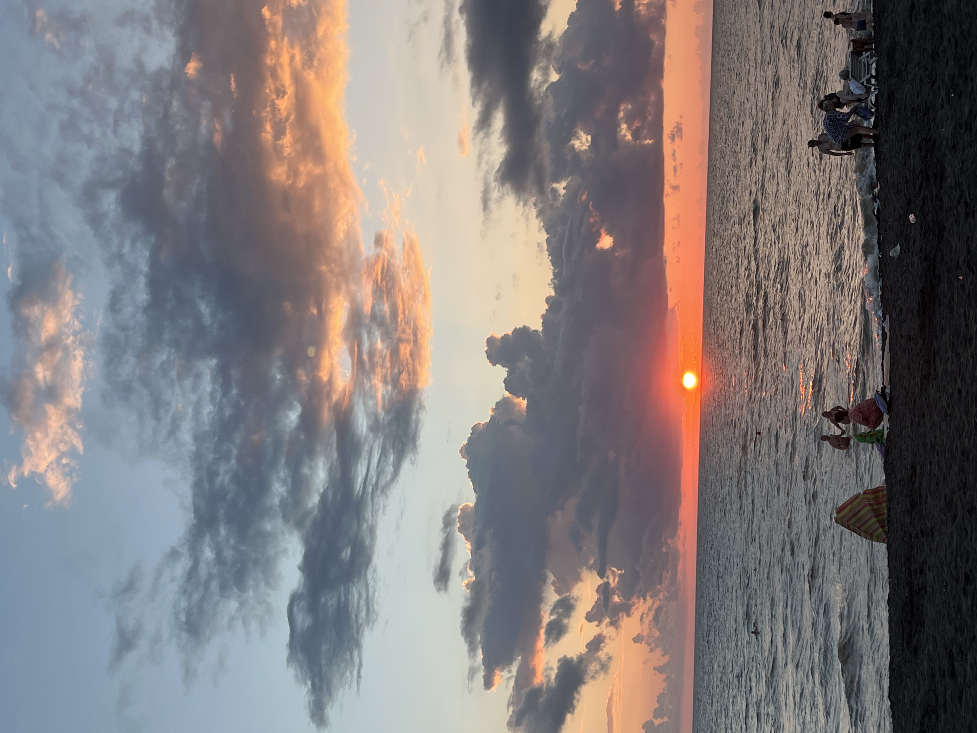 The image depicts a sunset at Ureki Beach, Georgia, a location known for its magnetic black sand. The sky is filled with dramatic clouds, tinged with oranges, pinks, and grays, creating a vibrant backdrop for the setting sun, which appears as a bright, orange orb just above the horizon. The sea is calm with gentle waves lapping at the shore.
On the dark, sandy beach, several people are visible. In the foreground, a colorful striped umbrella is set up, with a few individuals gathered around it. Further down the beach, closer to the water, groups of people are standing, some interacting, while others are wading in the shallow sea. A few individuals are also in the water further out. The overall scene is one of a relaxed evening at the beach, with the natural beauty of the sunset providing a picturesque setting.