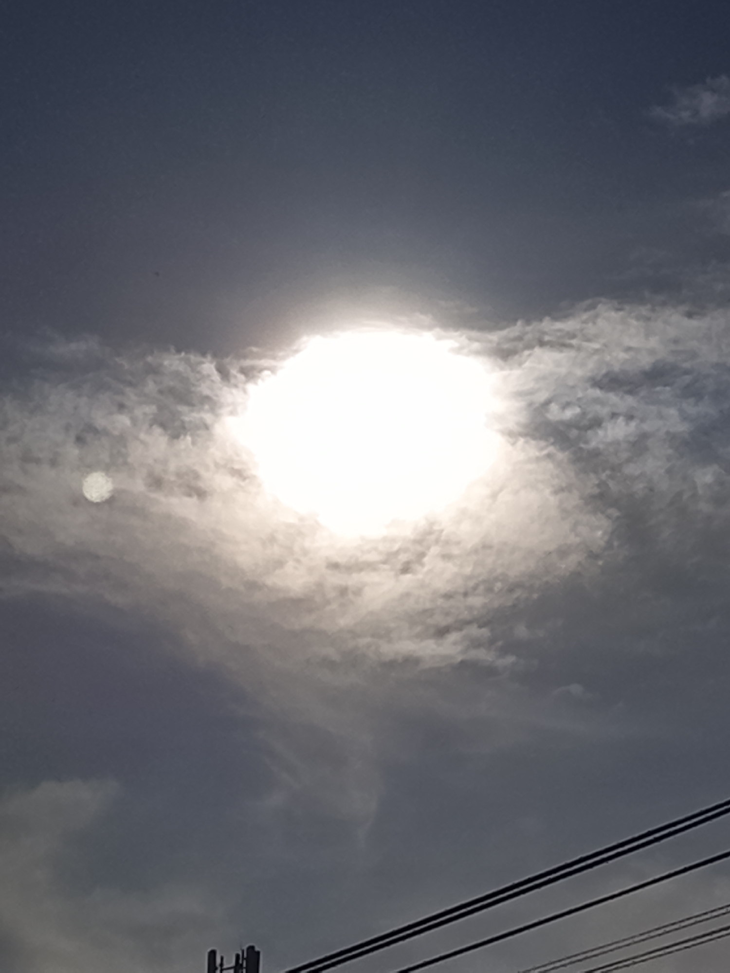 A bright sun is visible in a cloudy sky, with a hazy halo around it. To the left of the sun, a smaller, circular light object is also present. In the foreground, power lines and a cell tower are visible against the dark sky. The image appears to be taken in Lhokseumawe, Indonesia, possibly during daylight hours, with the sun partially obscured by clouds.