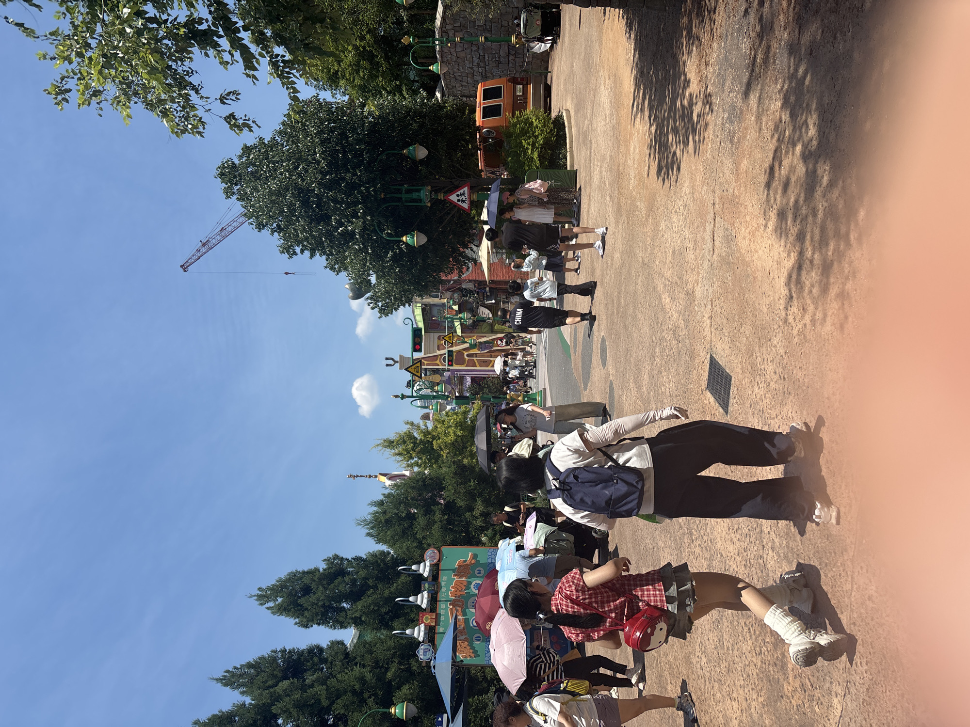 This is a wide-angle, eye-level outdoor shot of a public space, likely a theme park, in Pudong, China. The scene is bathed in bright sunlight, casting dappled shadows from overhead foliage onto the reddish-brown paved ground. In the mid-ground, several people are walking, some holding umbrellas to shield themselves from the sun. Two young women are prominent in the foreground, walking away from the camera. The woman on the left wears a red and white plaid shirt, a denim skirt, and leg warmers. She carries a red Hello Kitty-themed backpack. The woman to her right has a blue backpack and wears a white long-sleeved shirt and dark pants. To their right, a family with a child in a stroller is visible. In the background, there are various structures with colorful, cartoonish architecture, suggesting an amusement park setting. Trees line the path and taller evergreen trees frame the left side of the image. A red construction crane is visible in the distance against a clear blue sky. An orange van is parked on the right side of the path. The overall atmosphere appears to be one of a busy, sunny day in a recreational area.