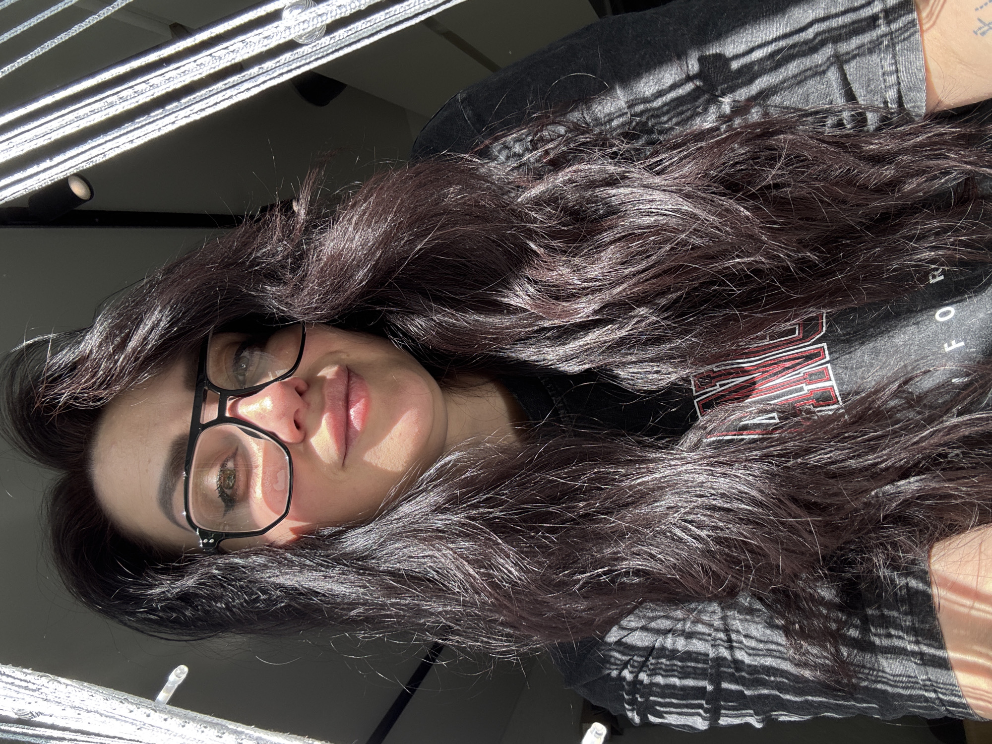 A medium-shot, rotated portrait captures a woman with dark hair and eyeglasses. Her long, wavy hair is voluminous and frames her face. She is wearing glasses with thick, black frames. The lighting is bright, with harsh shadows cast across the scene, likely due to sunlight filtering through blinds. The woman is wearing a dark-colored t-shirt. The location is implied to be indoors, based on the presence of blinds and lighting fixtures in the background. The setting and the city are not specified.
