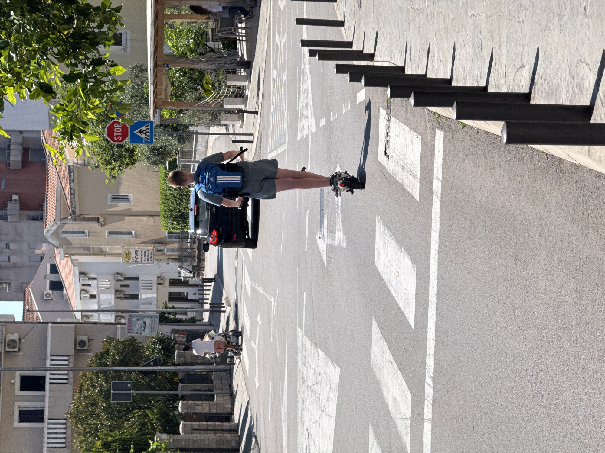A young Caucasian male wearing a blue backpack and grey shorts is riding an electric scooter across a pedestrian crossing. He is facing away from the camera, moving from left to right. In the background, a dark car is stopped at a traffic light, and a person is pushing a stroller. Buildings line the street, with trees and foliage interspersed. A stop sign and a pedestrian crossing sign are visible on a pole. The scene appears to be in a city, likely Budva, Montenegro, based on the architectural style and streetscape.