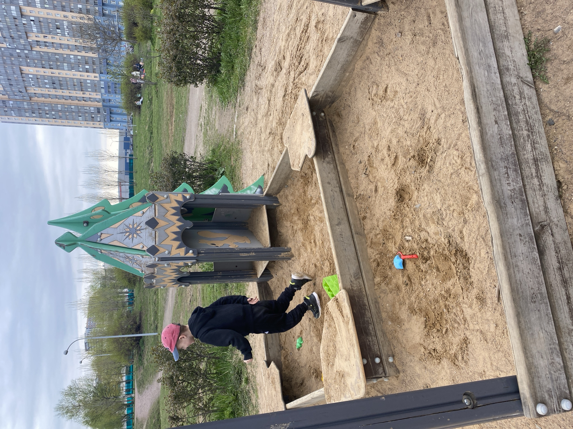 A color photograph shows a young child playing in a sandbox. The child is wearing a black outfit with a red and blue baseball cap, and black and gray sneakers. The child appears to be kneeling or crouching in the sandbox.
The sandbox is constructed of light-colored wooden planks. Inside the sandbox are a few colorful plastic toys, including a red and blue shovel.
To the left of the sandbox is a green and gray play structure that resembles a small castle or tower. The structure has decorative designs and shapes cut into its sides.
The sandbox is located in a park or playground, with grass and bushes visible in the background. In the distance is a tall building with many windows. The sky is overcast and gray.
Based on visual clues and publicly available information, the image may be from Saint Petersburg, Russia.
