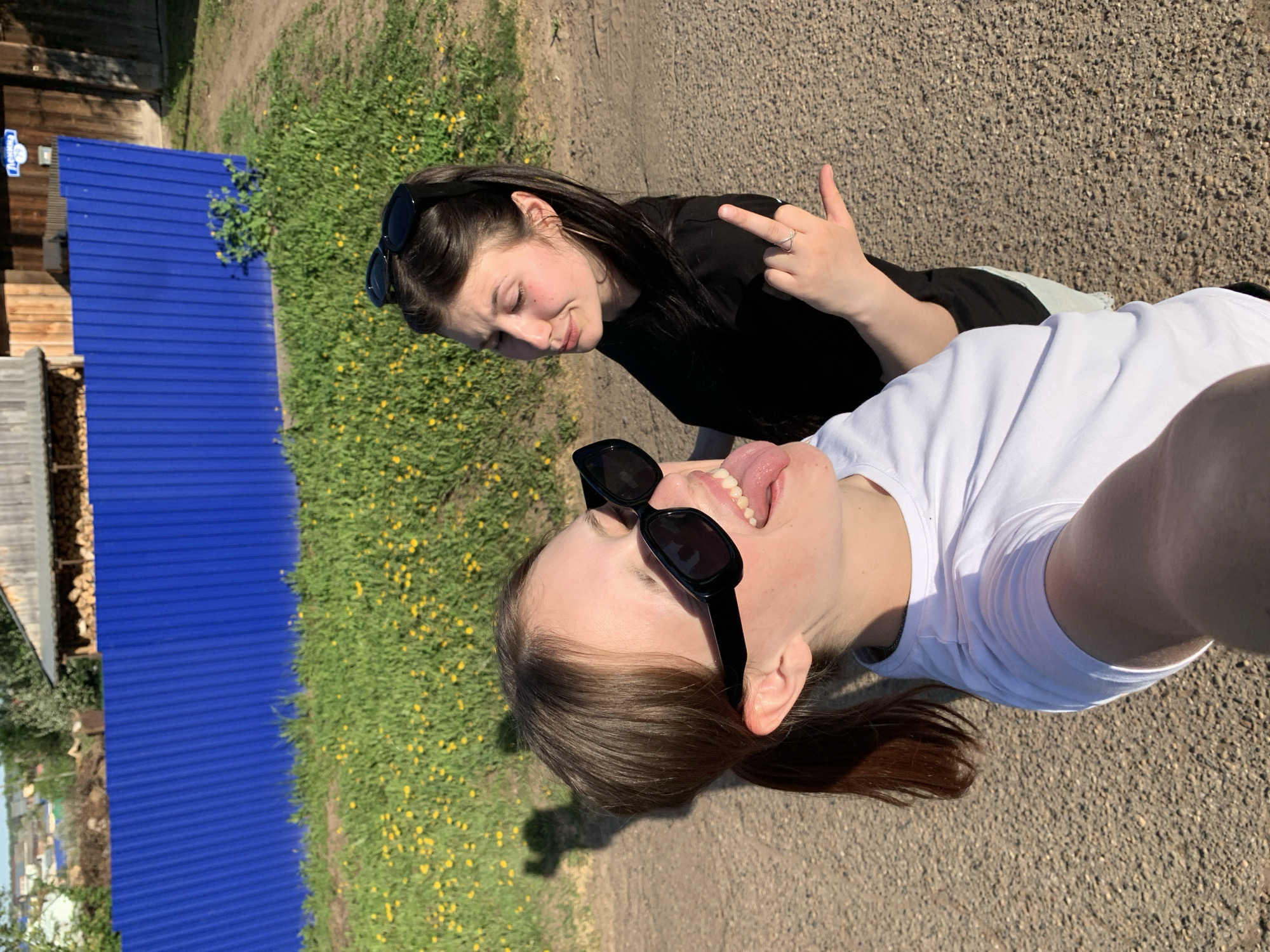 Two women pose for a selfie in Фоки, Russia. One woman, wearing a white t-shirt and sunglasses, sticks her tongue out. The other woman, wearing a black t-shirt and sunglasses, displays her middle finger. A blue metal fence is visible in the background, along with a gravel path and vegetation with yellow flowers. A wooden structure is also visible.