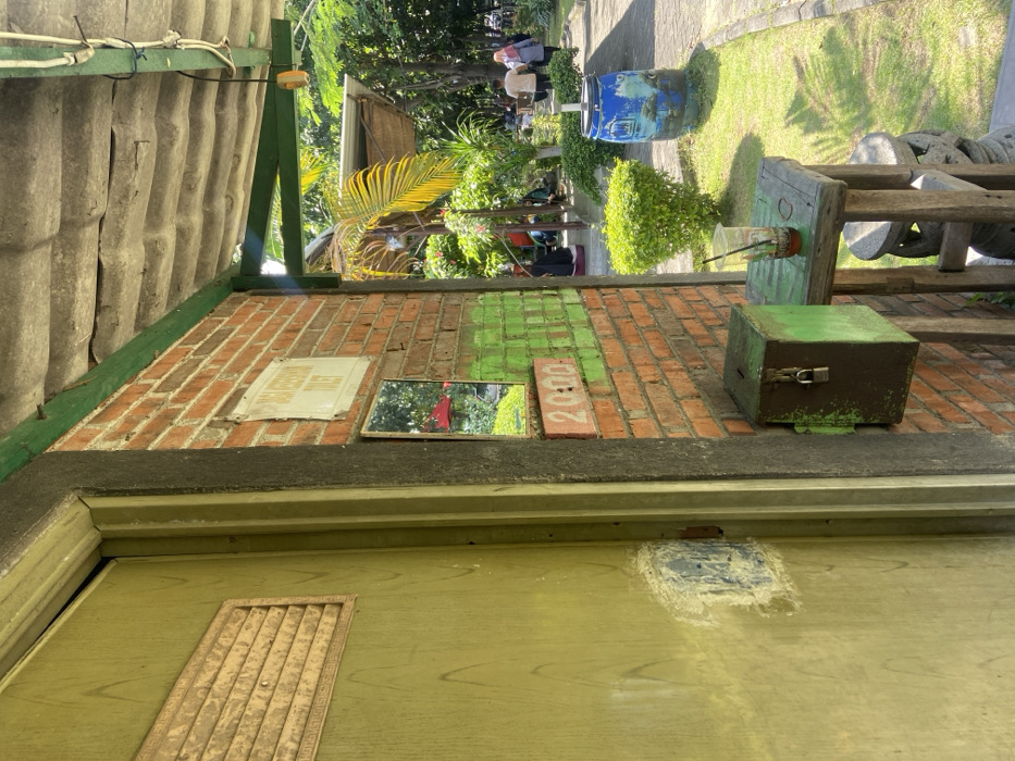 The image shows a brick wall with a green door to the left and a green wooden box to the right. The door is marked with a sign that reads “Toilet”. The box is open and appears to be a donation box with a lock. On the wall is a mirror and a painted number “2000”. The scene is located in Kecamatan Sidoarjo, Indonesia, at coordinates -7.442, 112.705. In the background, a group of people walk away from the scene on a paved pathway, through a lush, green lawn and a small garden. There is a blue barrel in the background. A wooden table with a glass of drink sits in the foreground. The table is made of unfinished wood and has two shelves. There is a small wooden stool in the lower right corner of the image. A long green roof overhangs the scene. The scene is bright and sunny, with the sky mostly obscured by the roof. The overall impression is of a place of simple beauty and quiet contemplation.
