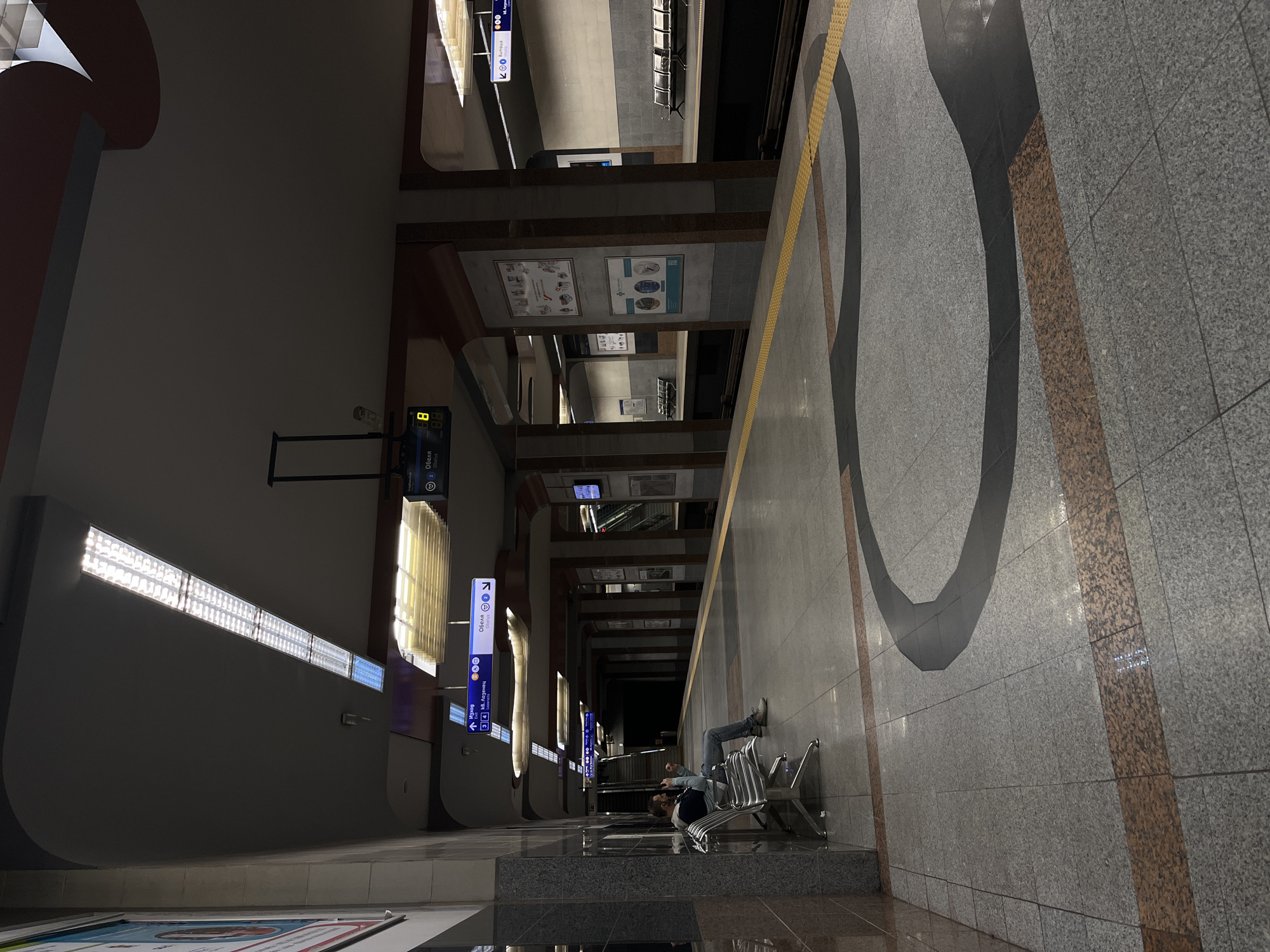 The image depicts the interior of a subway station, potentially in an Eastern European city, given the Cyrillic script on the signs. A person is seated on a metal bench, partially obscured by the angle of the shot, appearing to be waiting or resting. The station platform is wide and tiled with a combination of grey and brown, with a distinctive black curved design on the floor. A yellow line runs along the edge of the platform. Overhead, there are rows of fluorescent lights and direction signs, indicating "Изход" (Exit) and "Обеля." The perspective suggests the photo was taken from an elevated position, looking down the length of the platform towards the darkened tunnels where the train would arrive. The station appears modern with clean lines and a muted color palette.