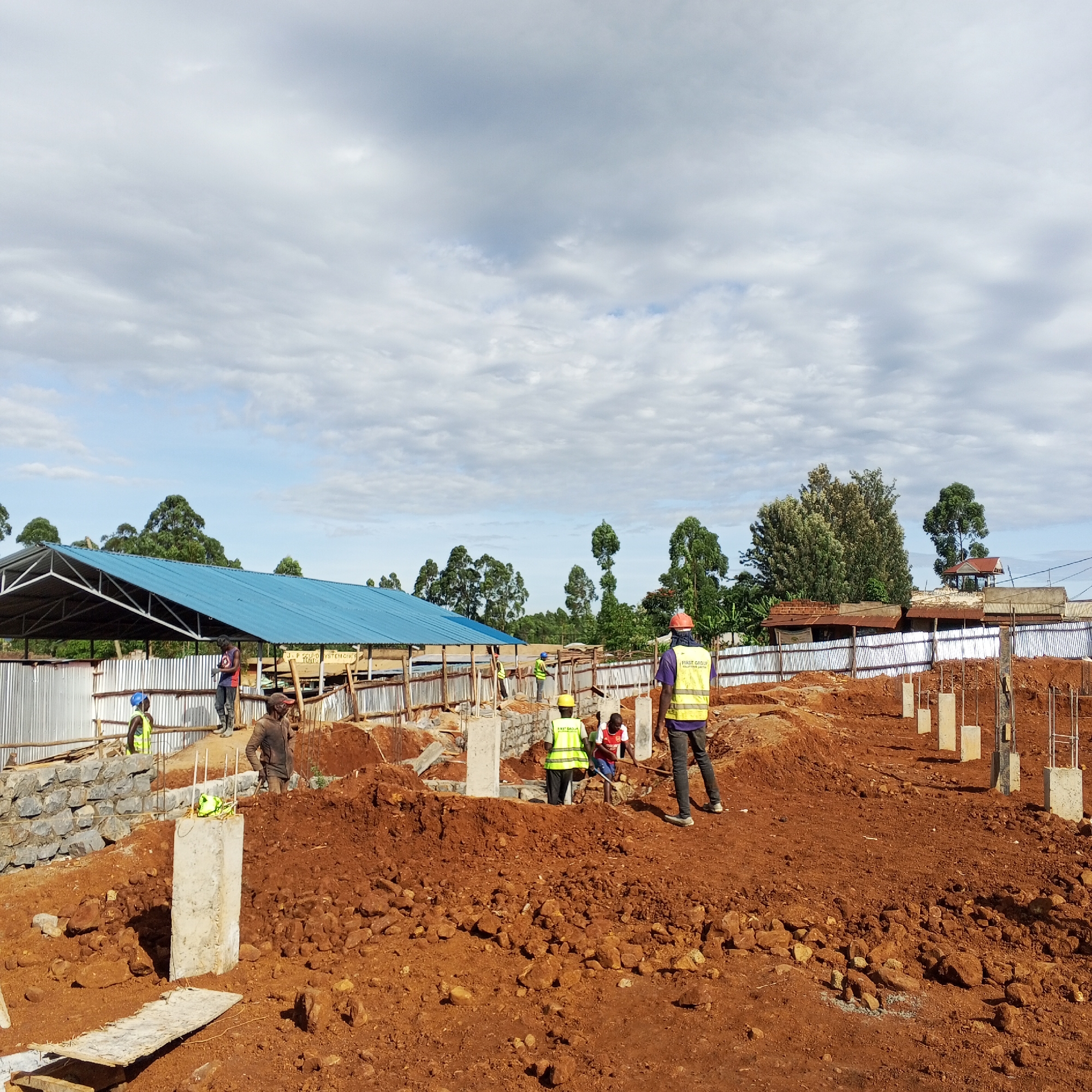 The image depicts a construction site in what appears to be the Borabu Chitago ward in Kenya. Red soil dominates the foreground and midground. Several concrete pillars and partially built structures are visible. The pillars have exposed rebar protruding from their tops. Construction workers, wearing safety vests and helmets, are spread throughout the site. Some are working on the foundation, while others stand and observe. In the background, a structure with a blue roof is apparent, along with fencing and trees. The sky is overcast, with a mix of blue sky and clouds.