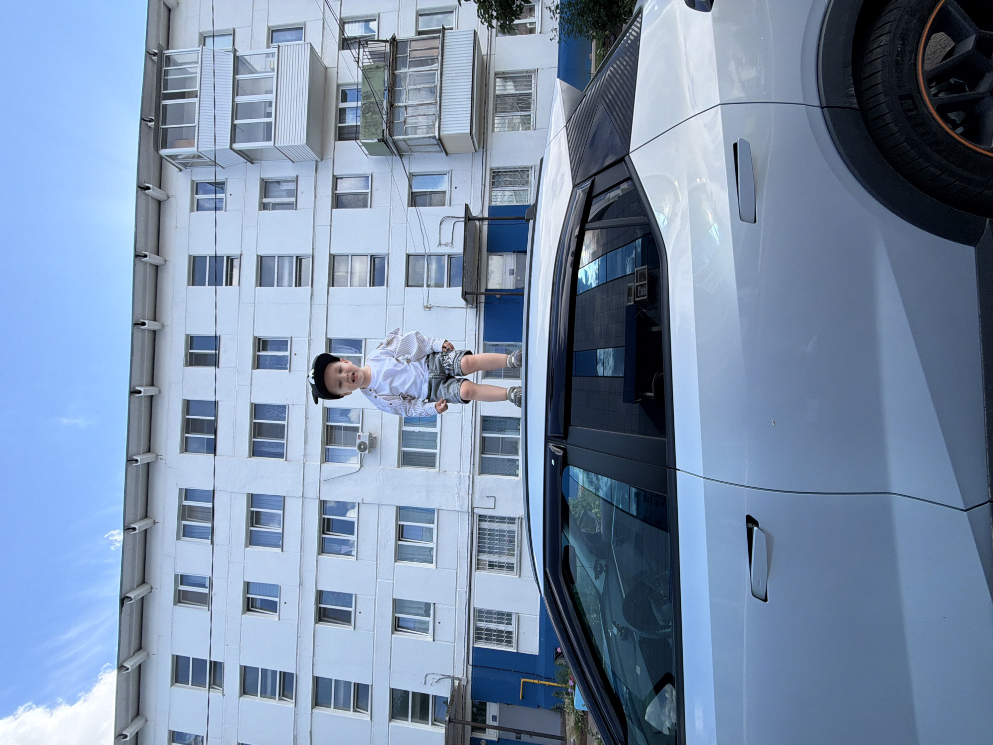 A young boy, wearing a black cap, white shirt, and grey shorts, stands on a white car. He is positioned on the roof of the car, with his legs stretched out and arms raised. The boy's mouth is open, suggesting he might be smiling or speaking. The car is parked in front of a tall, white apartment building with many windows. The building appears to be old, with some of its windows covered by metal grates. Some sections of the building are painted blue. The sky is blue with some scattered white clouds, indicating it is daytime and the weather is fair. A tree branch with green leaves is visible at the top right of the image, suggesting it might be spring or summer. The scene appears to be a residential area in Chaikovsky, Russia, given the architectural style of the building.