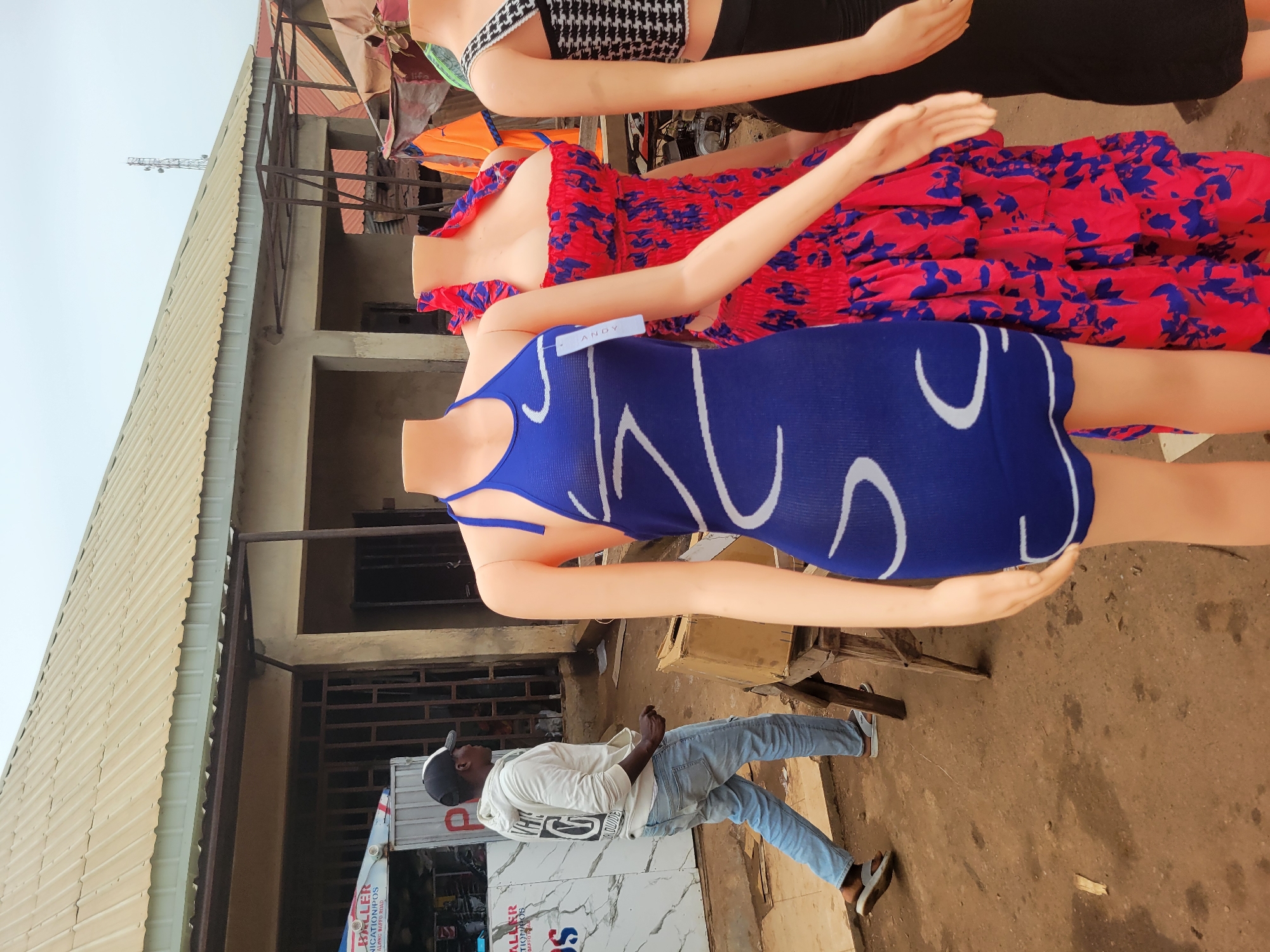 This image captures an outdoor market scene in Bokkos, Nigeria. In the foreground, three headless mannequins are displayed. The mannequin on the left wears a blue halter dress with white abstract patterns. The middle mannequin is adorned in a red and blue floral print dress with ruffled layers. The mannequin on the right is dressed in a black and white patterned top and a black skirt. Behind the mannequins, a man wearing a white hoodie, blue jeans, and a black and white cap walks away from the camera, his gaze directed to the right. He is wearing grey and white sandals. The market appears to be set up under a corrugated metal roof supported by metal poles. To the left of the mannequins, a stall is visible with various items displayed, including a cooler and signage. The ground is a mixture of dirt and concrete. The overall environment suggests a bustling, open-air marketplace typical of a Nigerian town.