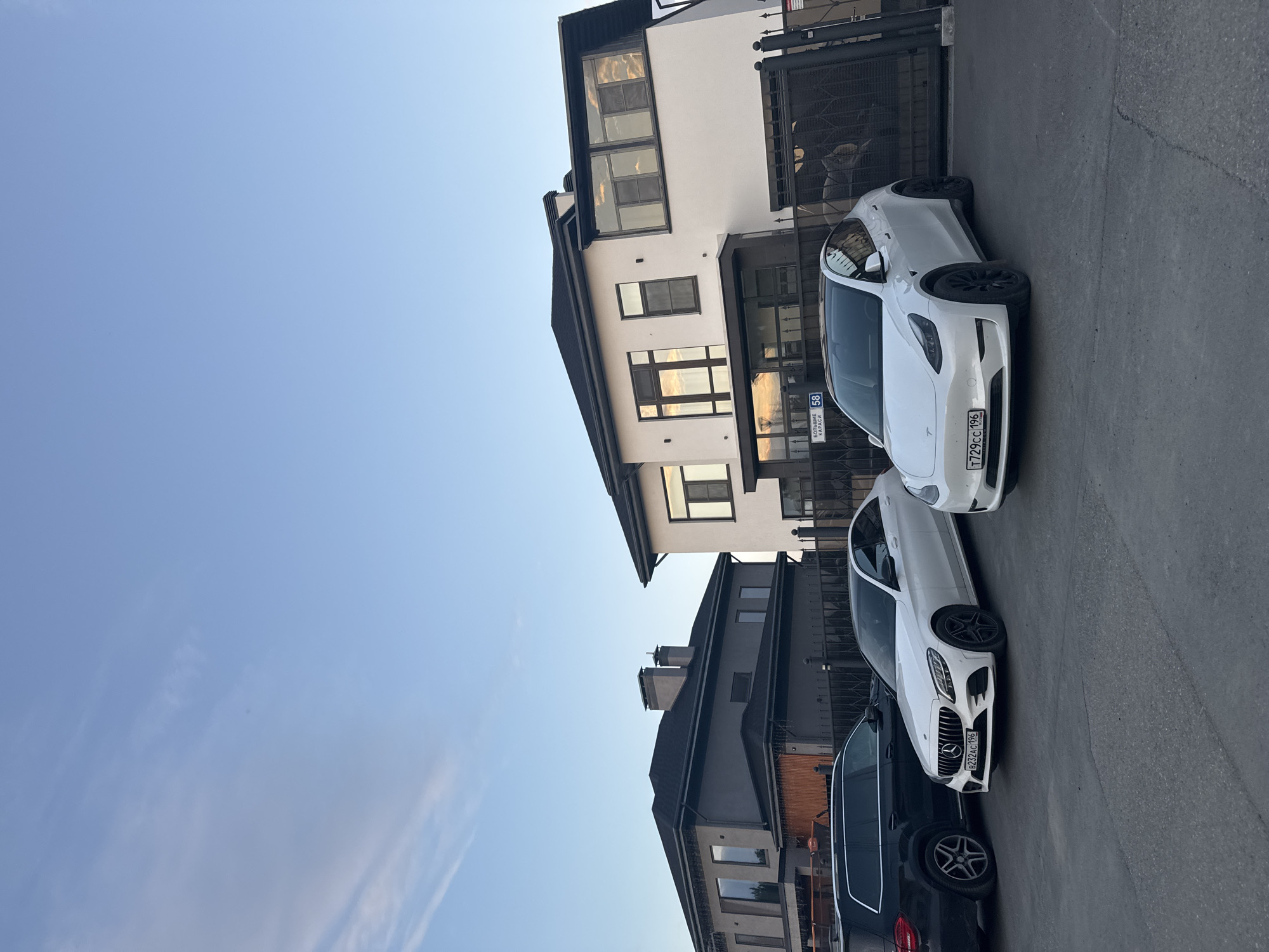 The image shows a street scene in Yekaterinburg, Russia. Two white cars and one black car are parked on an asphalt road in front of modern buildings with dark roofs and light-colored walls. The white cars appear to be luxury vehicles, one with a Tesla badge and the other a Mercedes-Benz, both with license plates that are visible. The Tesla's license plate reads "T729CC 196". The Mercedes-Benz has a license plate with Cyrillic characters and numbers. The buildings have large windows that reflect the sky. A metal fence is visible in the background, and a sign with "58" is also present. The sky is clear blue with a few wispy clouds, suggesting it is daytime. The overall impression is one of a well-maintained residential or commercial area. No people are visible in the image.