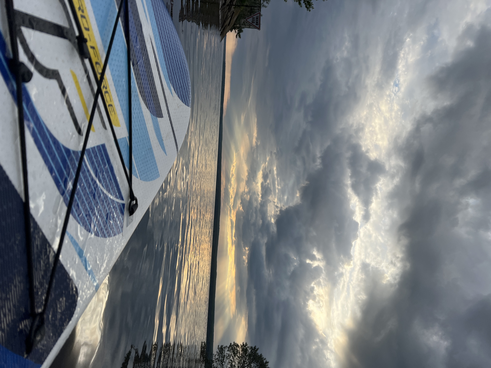 The image captures a serene waterscape from the perspective of a paddleboard (SUP), positioned in the foreground on the left. The white paddleboard features blue, black, and yellow designs, with the text "PRO FORCE" visible on its surface, held by black bungee cords.
The board rests on calm, rippling water that reflects the warm hues of the sky. The scene is set at either sunrise or sunset, as golden light pierces through a heavily clouded sky, creating dramatic light rays on the horizon. The sky is dominated by dark, voluminous clouds, with brighter, illuminated patches where the sun's light breaks through.
A distant, narrow shoreline defines the horizon, with some trees and foliage visible. On this distant shore, a triangular white sign with a red border and black Cyrillic text is visible. The text reads "ТИХИЙ ХОД," which translates to "SLOW SPEED" or "QUIET RUN," indicating a specific regulation for the waterway. The setting evokes a peaceful yet majestic atmosphere. The location is Chaikovsky, Russia.