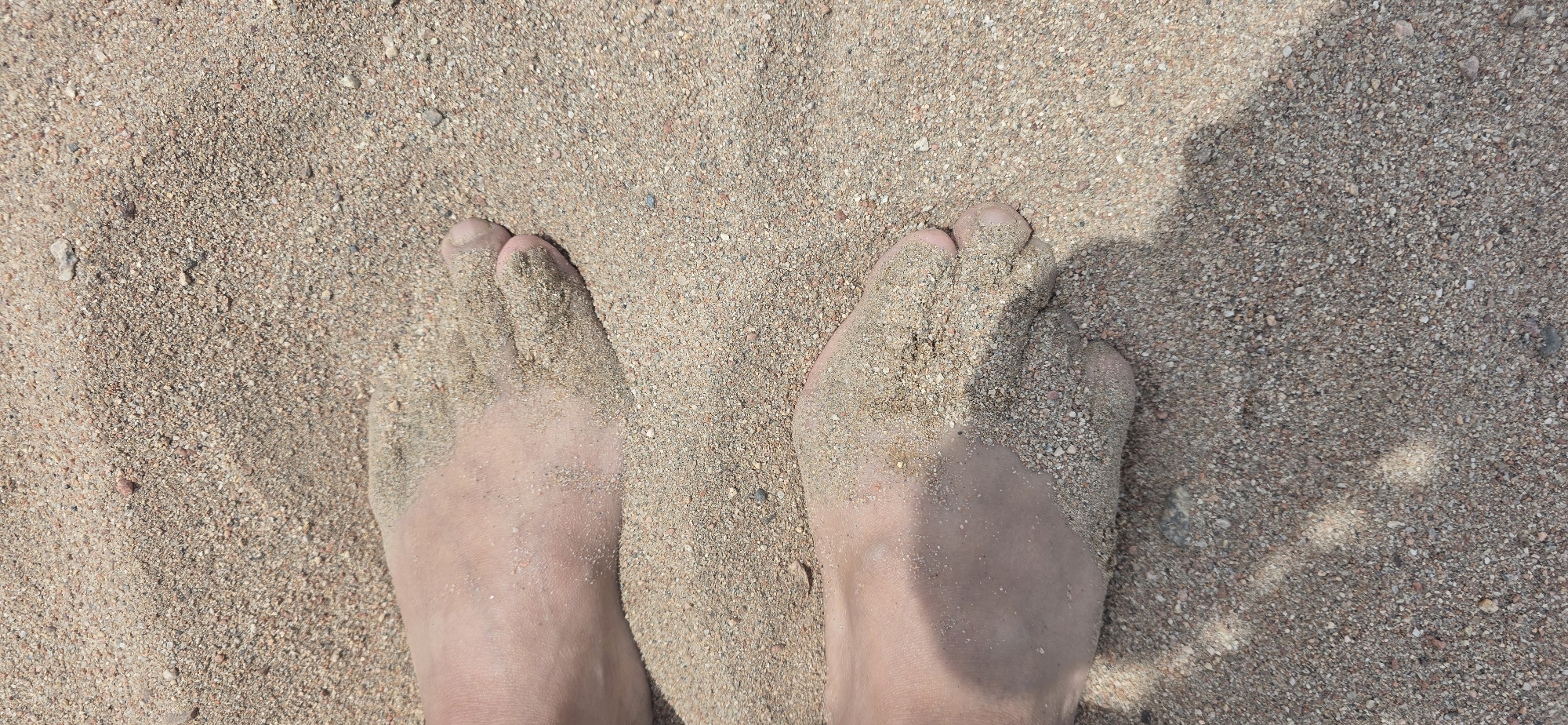 The image shows a pair of human feet partially buried in coarse, light-brown sand on a beach. The toes of both feet are visible, with sand adhering to the skin. The lighting suggests it is daytime, and a shadow is cast across the sand, indicating the presence of an unseen object or person above. The environment is a sandy beach, with the sand appearing to be a mix of fine grains and small pebbles.