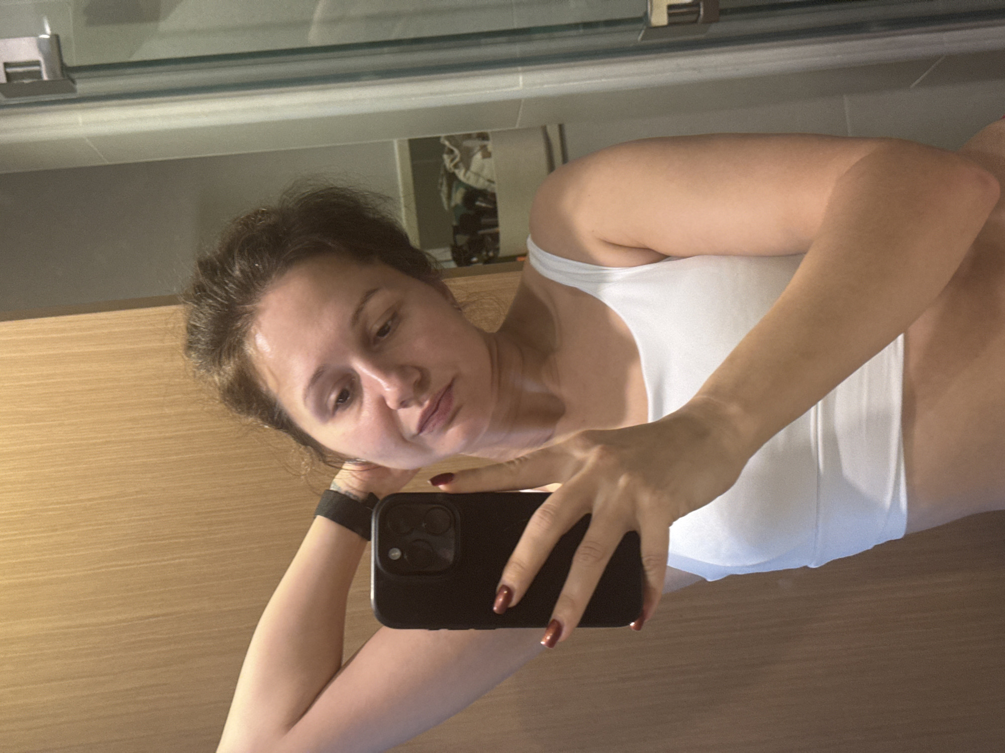 A woman takes a selfie in a bathroom. She holds a black smartphone, her face tilted toward the camera. She wears a white top and a black fitness tracker on her wrist. The bathroom features wood-paneled walls and a mirror reflecting a partially visible interior space. The location is Dubai, United Arab Emirates.