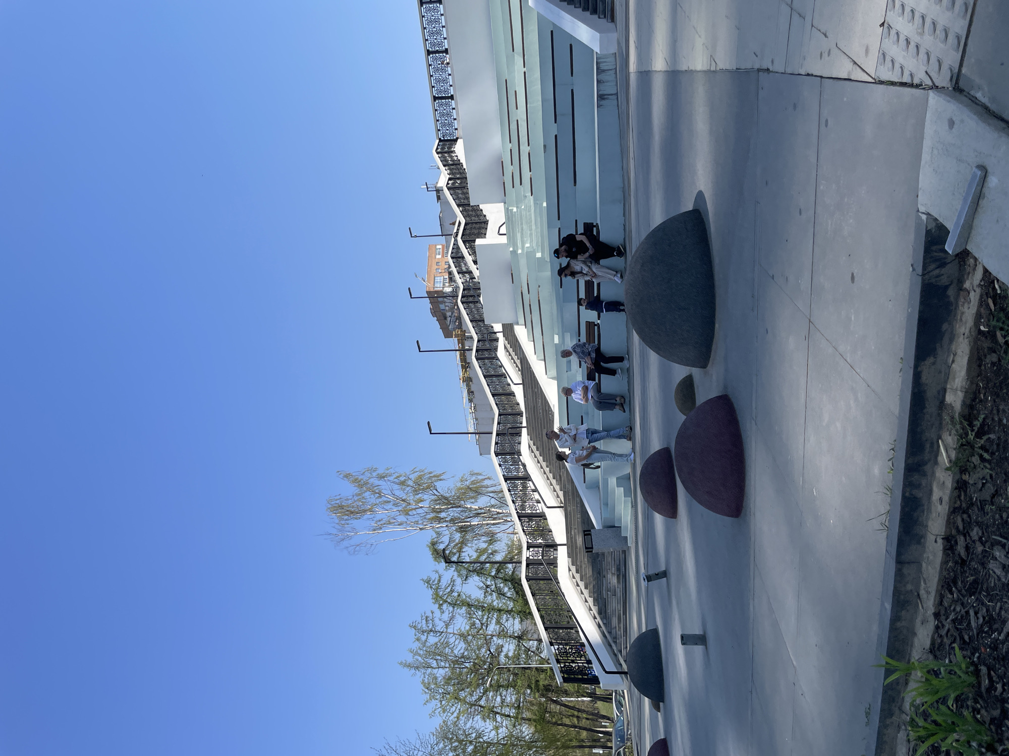 Here's a concise description of the image:
The image shows a terraced public space with people sitting on benches along a staircase structure. The staircase features decorative railings and white risers. There are modern, spherical seating elements along the walkway. Trees are visible along the side of the structure. The sky is clear and blue. The location is the central embankment of the Izhevsk pond in Izhevsk, Russia. People are sitting and relaxing on the staircases, enjoying the outdoor space.