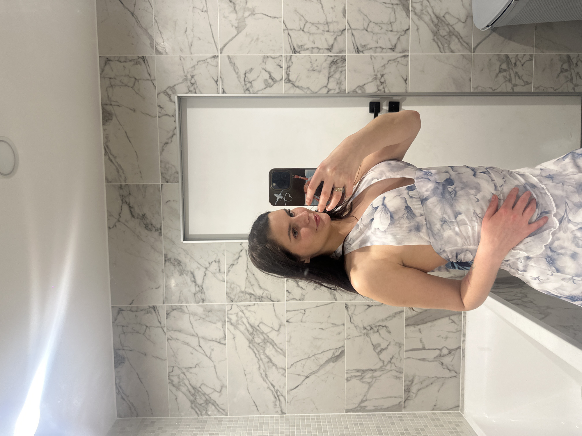 A woman stands in a bathroom, holding a phone and taking a mirror selfie. She has long, dark hair and is wearing a light-colored dress with a floral pattern. The bathroom features marble-patterned tiles and a white bathtub. The scene is well-lit, with bright overhead lighting.