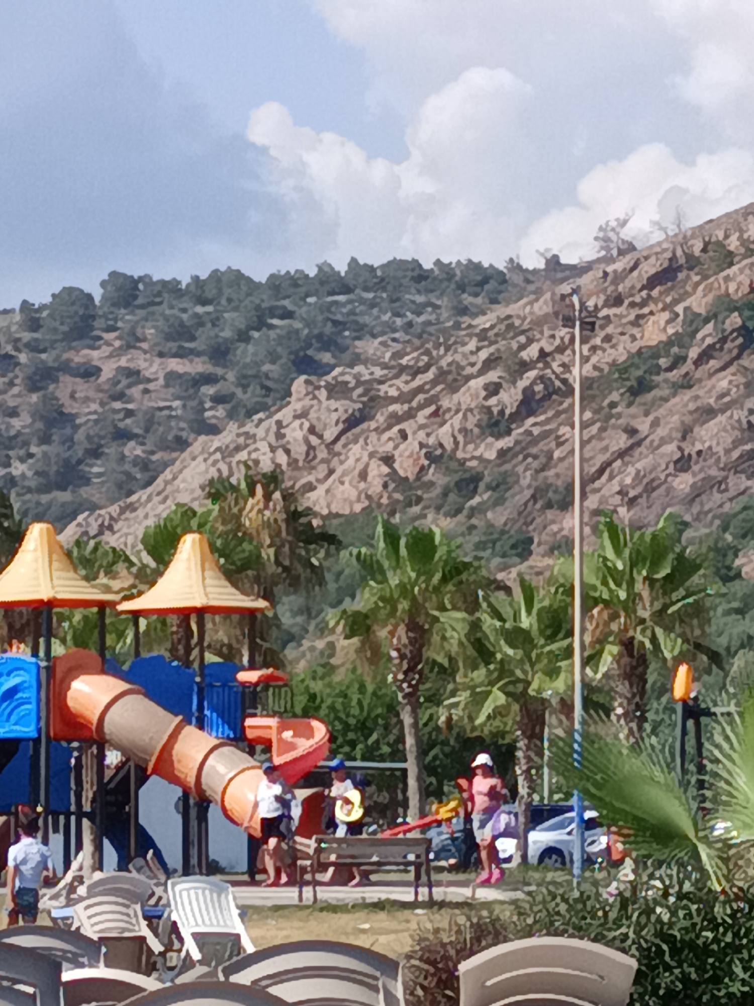 In Gazipaşa, Turkey, on a bright day, a children's playground is visible in the foreground. The playground features a large, multi-colored slide structure with orange and brown tubes, a red slide, and blue platforms. Several children and adults are present in and around the playground. One child in a white shirt and dark shorts stands near the bottom of the slide. Another child, wearing a white shirt and dark shorts, is walking away from the playground on the left. A woman in a pink shirt and gray shorts is standing near a small red slide on the right. An adult in a white shirt and dark shorts is standing on the playground. Behind the playground, palm trees and other greenery are abundant, with a rocky, tree-covered hillside forming the backdrop. The sky is blue with scattered white clouds. In the far right, a white car is parked. A metal light pole with an orange light fixture stands in the middle ground. Several empty white lounge chairs are scattered on the grass in the foreground.