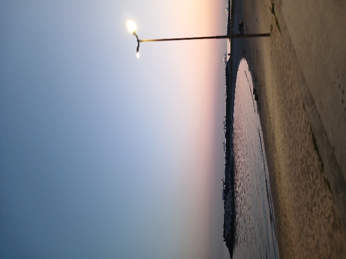 The image depicts a view of the shoreline at sunset from the Florya Beach in İstanbul, Türkiye, located at coordinates 40.956, 28.816. The photo was taken from the sandy beach, looking out towards the sea. In the distance, the seawall juts out into the water, forming a small bay. The water is calm and reflective, reflecting the pink and orange hues of the setting sun. A single streetlight stands tall on the right side of the image, its light illuminating the sand. Several people are gathered in the distance, seemingly enjoying the sunset on the beach.