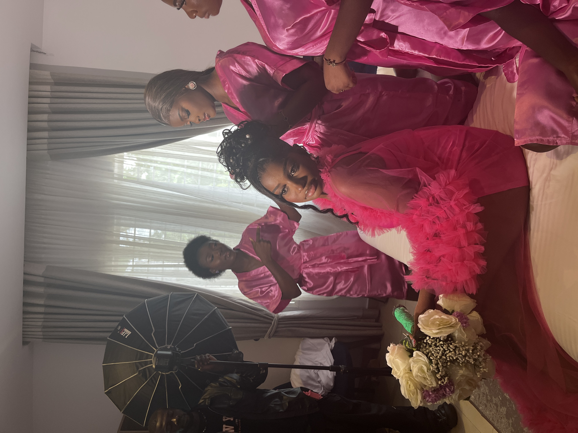 The image shows at least four women in matching pink satin robes posing for a photo indoors. One woman is holding a bouquet of white flowers. A photographer with studio lighting equipment is also present. The location appears to be a room with curtains in Gura Top, Nigeria.