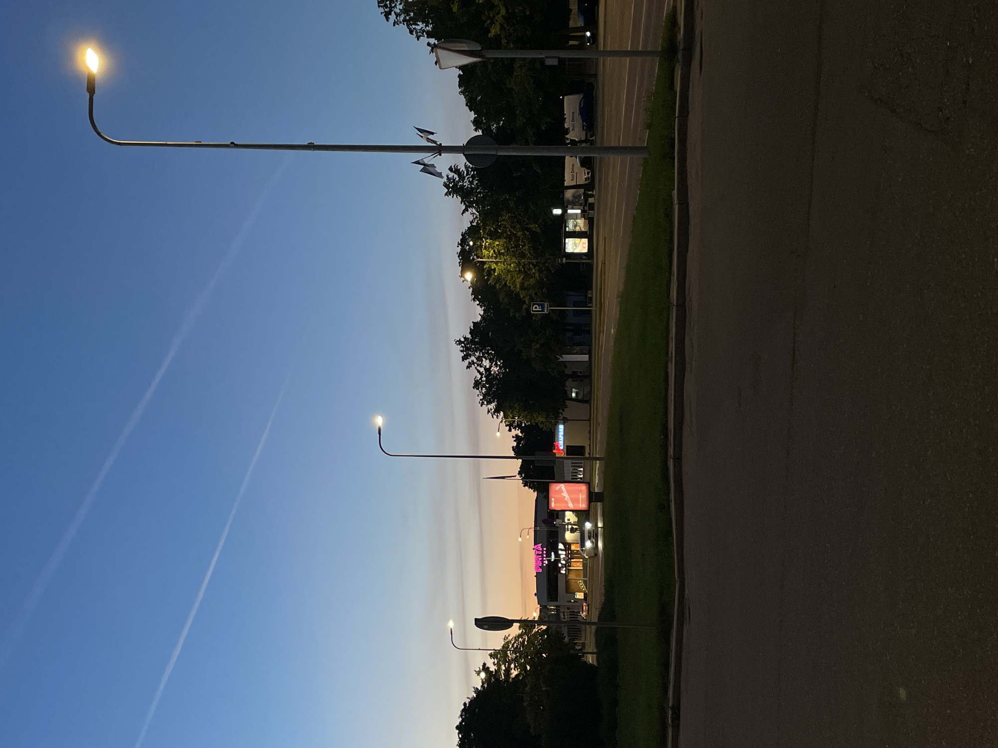 The image captures a street scene in Tallinn, Estonia, during twilight. The sky exhibits a gradient of colors, transitioning from a deep blue at the top to lighter shades of orange and pink near the horizon, with two contrails visible. Several streetlights are illuminated, casting a warm glow.
In the mid-ground, a building with illuminated signage that reads "PIRITA" in bright pink neon letters is visible. In front of this building, a white car is driving. Adjacent to the "PIRITA" building, to its right, are several digital billboards.
To the right of the "PIRITA" building and billboards, a row of dense, dark green trees forms a line along the edge of the road. Beyond these trees, more buildings and trees are faintly visible.
The foreground shows a paved road, with a grassy median strip to the left, demarcated by a curb. On the grassy median, a bus stop shelter with advertisements and a blue parking sign are present. Several vehicles, including a white van with "Bolt Drive" branding, are parked or moving on the road in the mid-ground and background. The scene is one of an urban environment at dusk, with artificial lights beginning to dominate the natural light.
