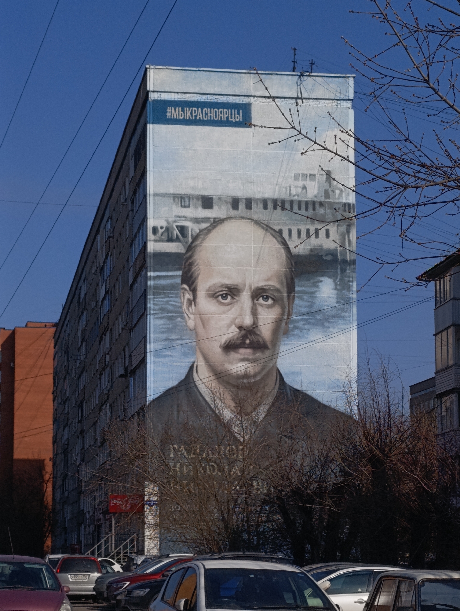 The image shows a large mural on the side of an apartment building. The mural depicts a portrait of a man with a mustache, wearing a dark suit. Above the man's head is a faded image of a boat on water. A blue banner runs across the top of the mural with text written in what appears to be Russian: #МЫКРАСНОЯРЦЫ. The building is likely located in the city of Krasnoyarsk, Russia. Several cars are parked in front of the building, and trees with bare branches are obscuring the lower portion of the mural. Other buildings are visible in the background under a clear blue sky.