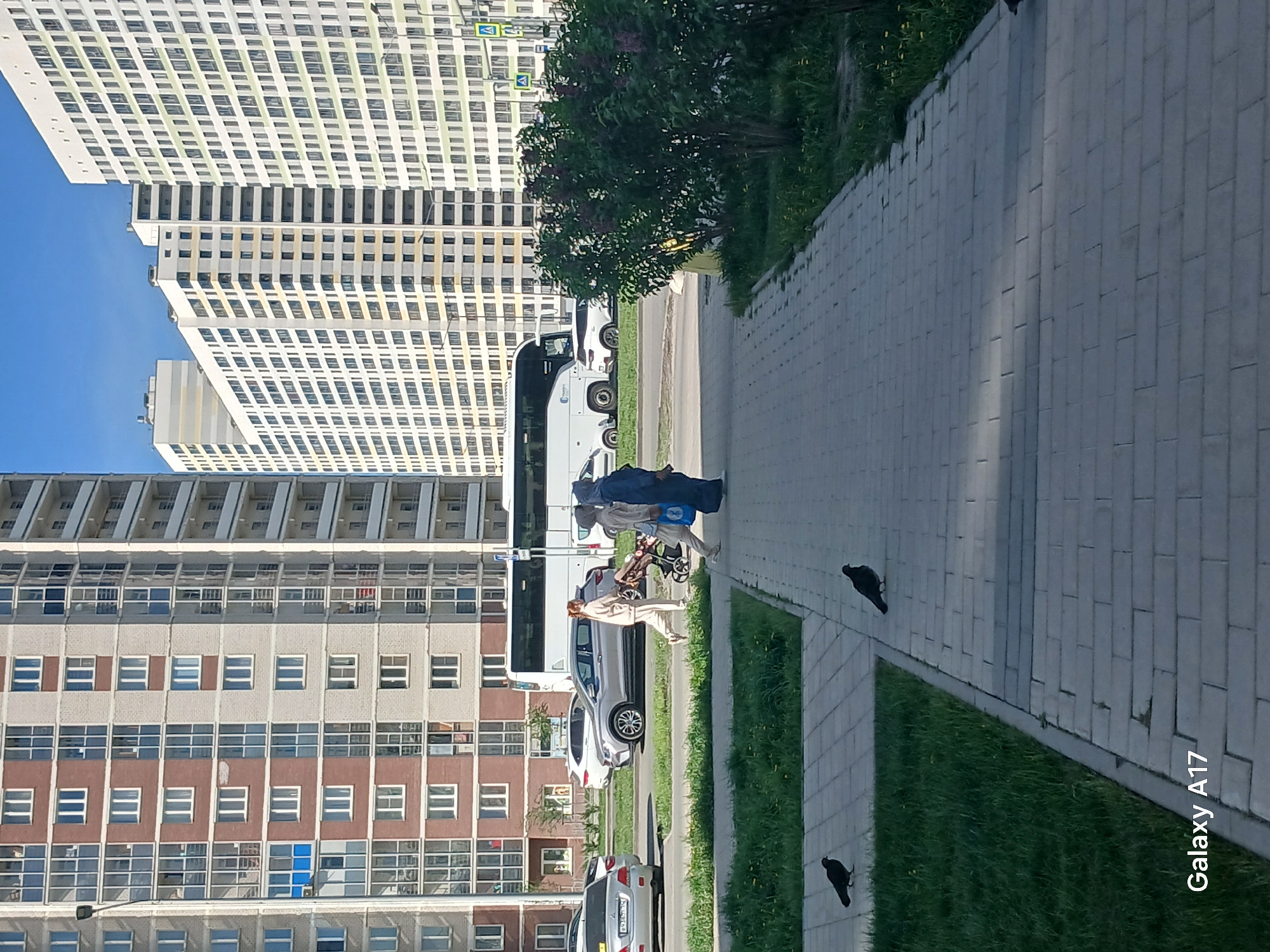The image depicts a street scene in Yekaterinburg, Russia. Several apartment buildings of varying heights and colors (white, grey, brown) line the left side of the frame. A paved sidewalk runs along the right side, bordered by a strip of grass. Two people, one dressed in a tan outfit and the other in a blue hijab, walk along the sidewalk, pushing a stroller. A white bus and several cars are parked along the road beside the sidewalk. Two black birds are visible on the sidewalk. The sky is blue.